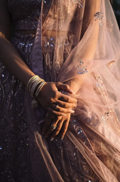 Close-up of a woman's hands gently resting on her lap, adorned with multiple bangles and rings, wearing a traditional embroidered dress with a sheer pink dupatta.