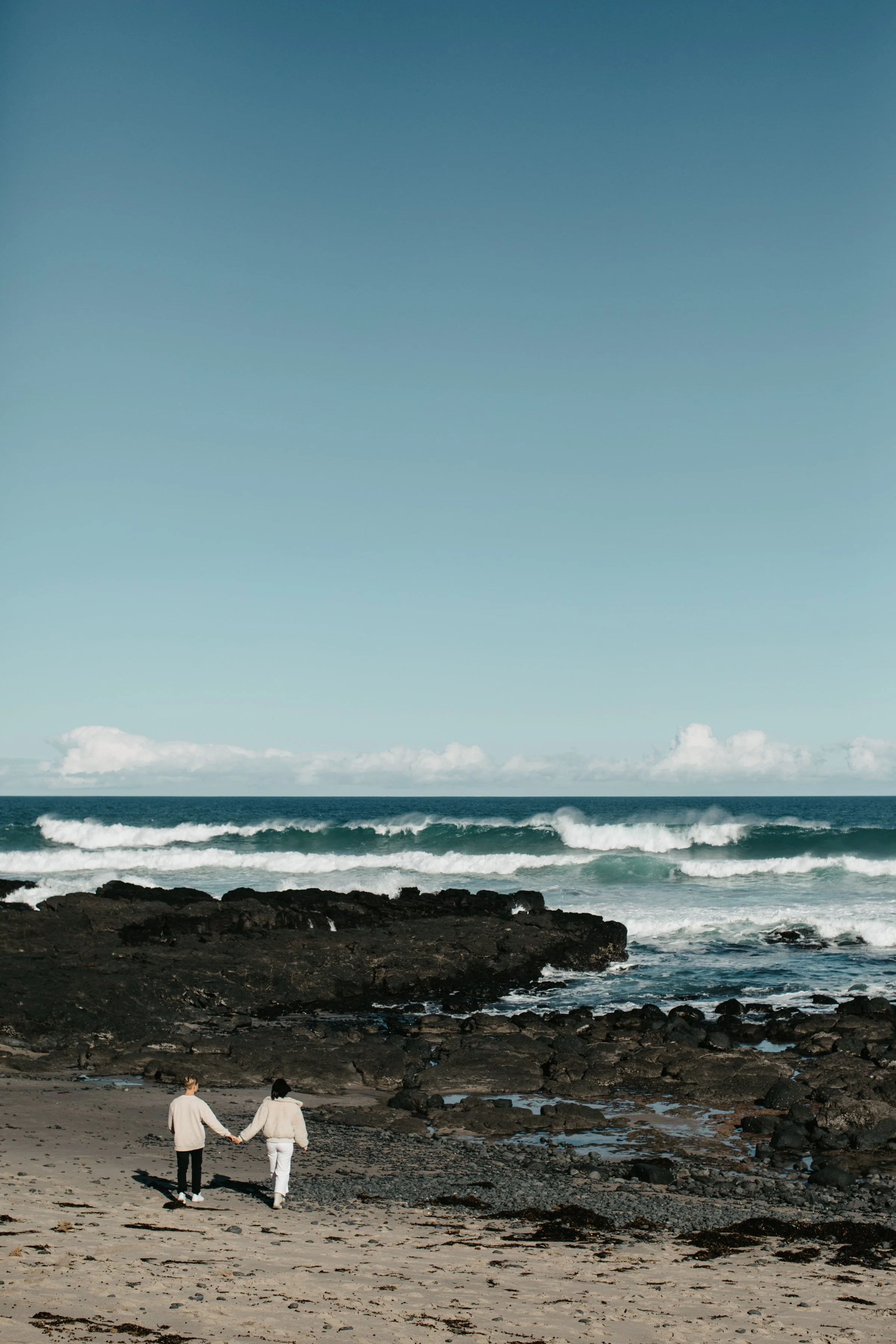 A couple holding hands and walking along a rocky beach shoreline with ocean waves and distant sky in the background.