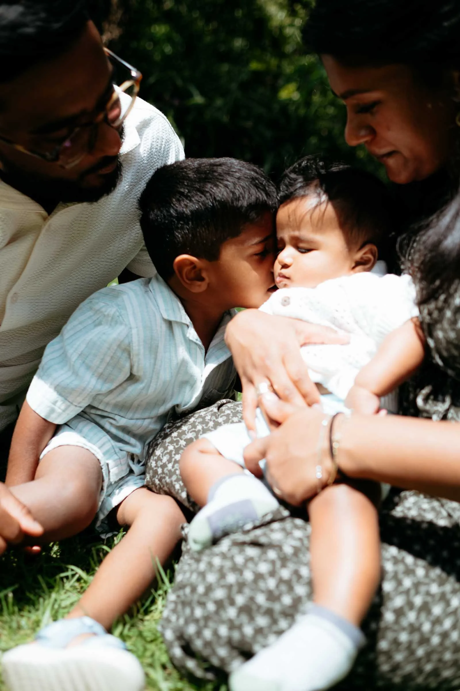 Two children, an older boy and girl, holding a baby girl outdoors, surrounded by greenery, in a warm, tender moment.