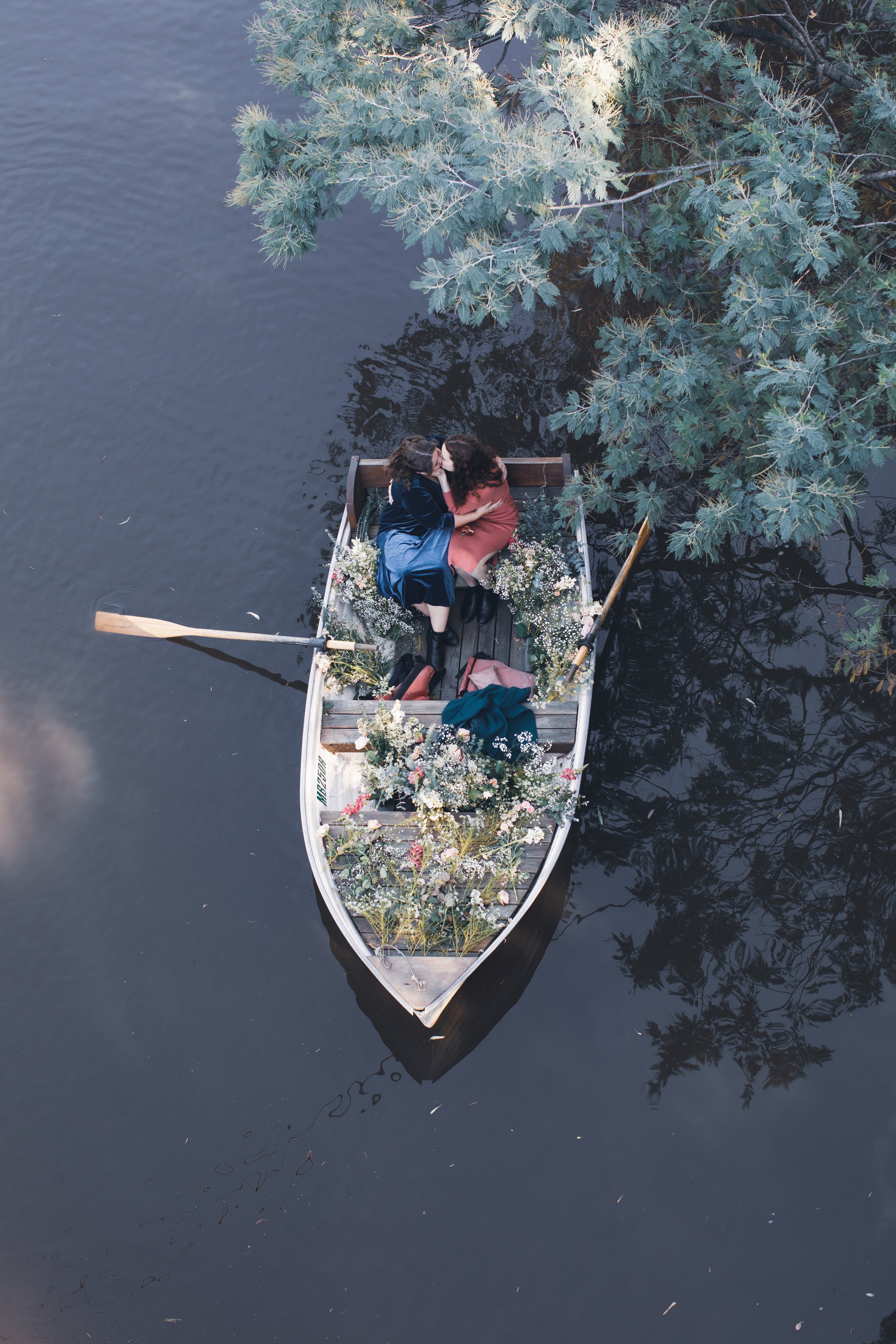 Two women are sitting in a small boat filled with flowers, kissing each other, on a dark river with overhanging tree branches.