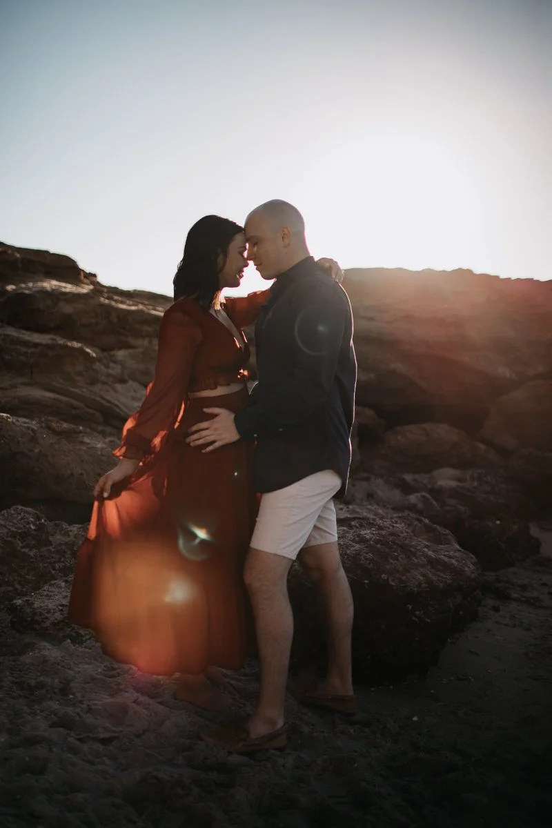 A couple embracing on a rocky beach at sunset, facing each other closely with their foreheads touching.