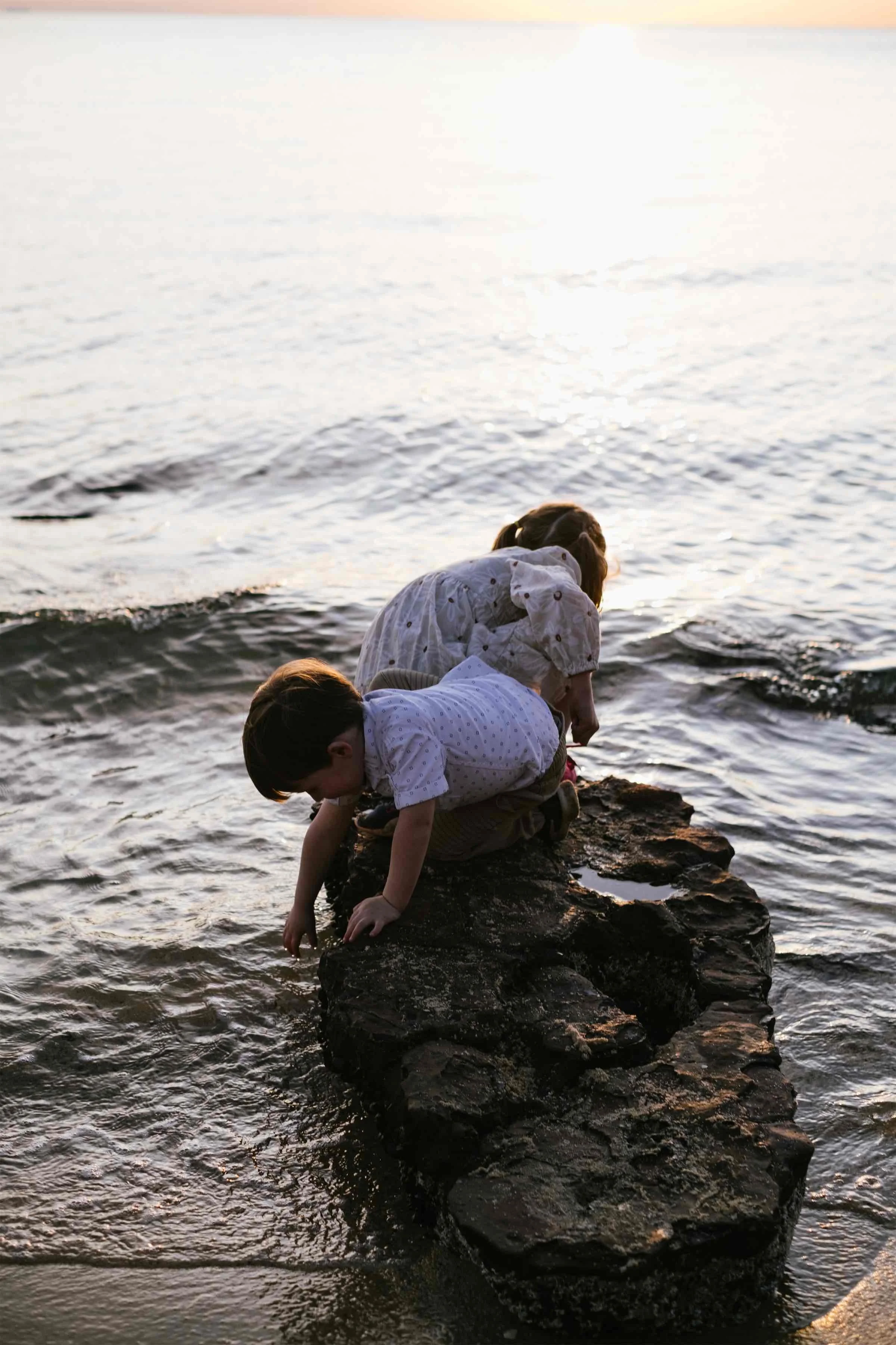 Two children, a girl and a boy, climbing on a rock by the water at sunset.