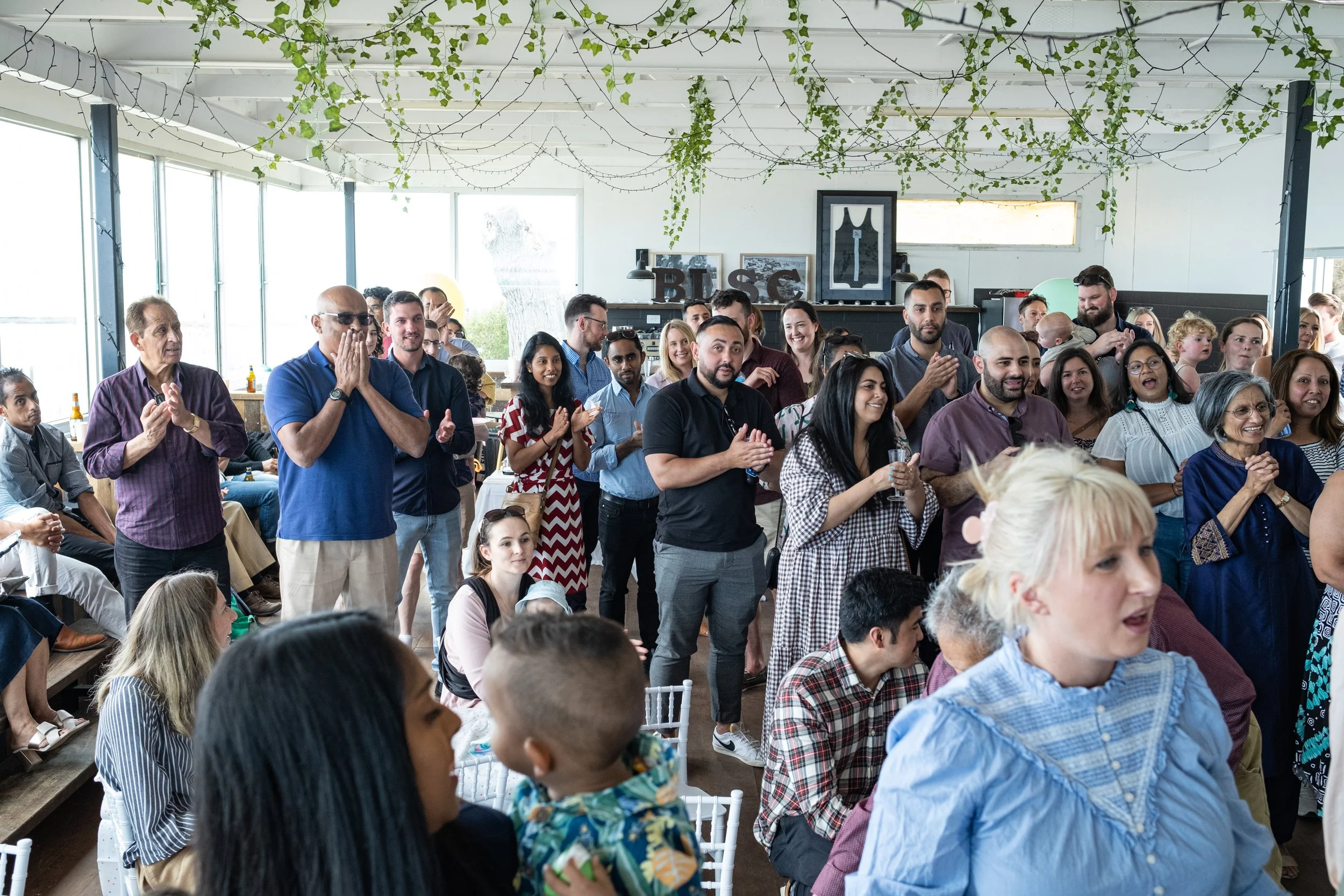 A large group of diverse people standing, clapping, and smiling at an indoor event with natural light, hanging green plants, and framed wall art.