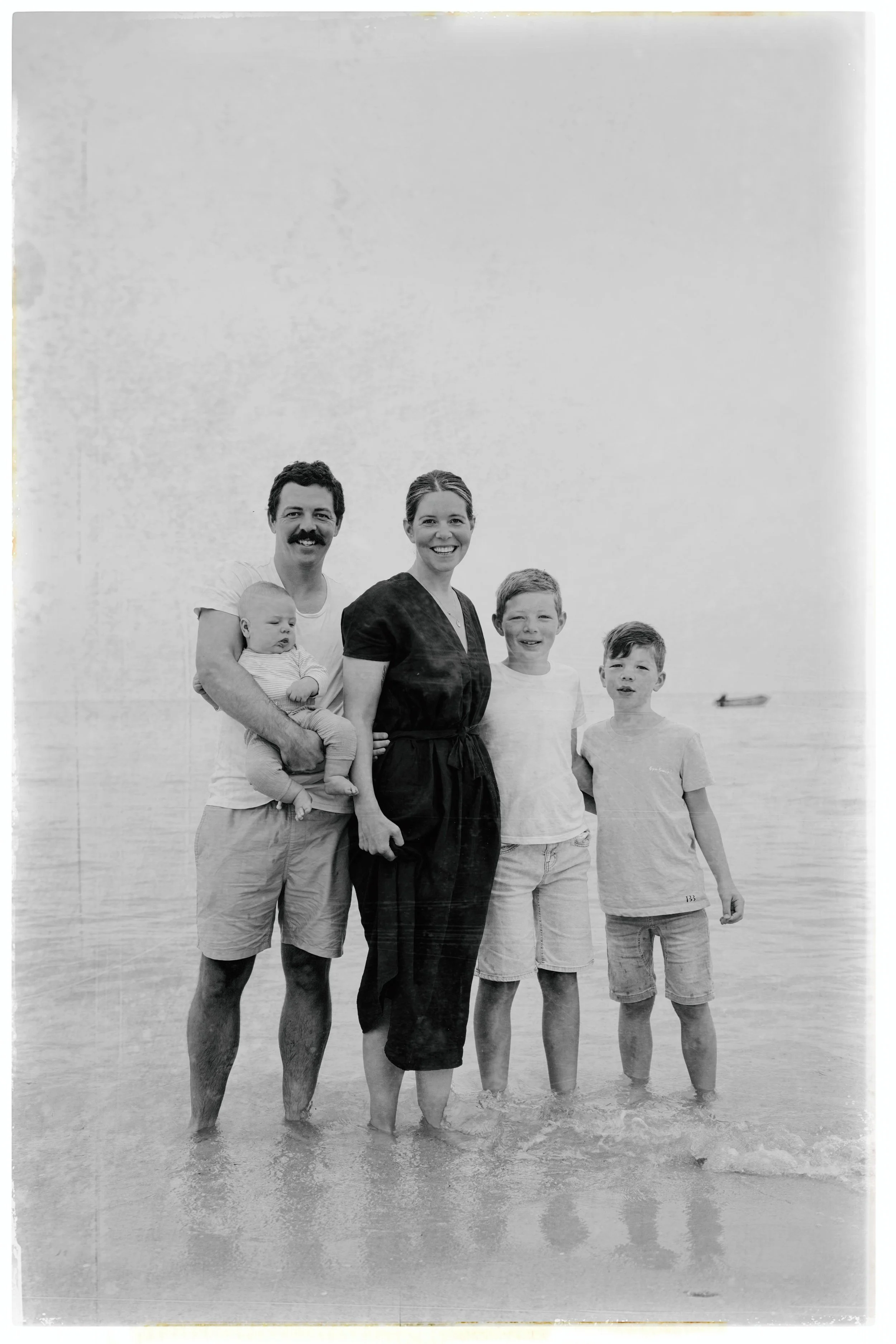 A family of five standing in the water at the beach, smiling and looking at the camera, with a boat in the background.