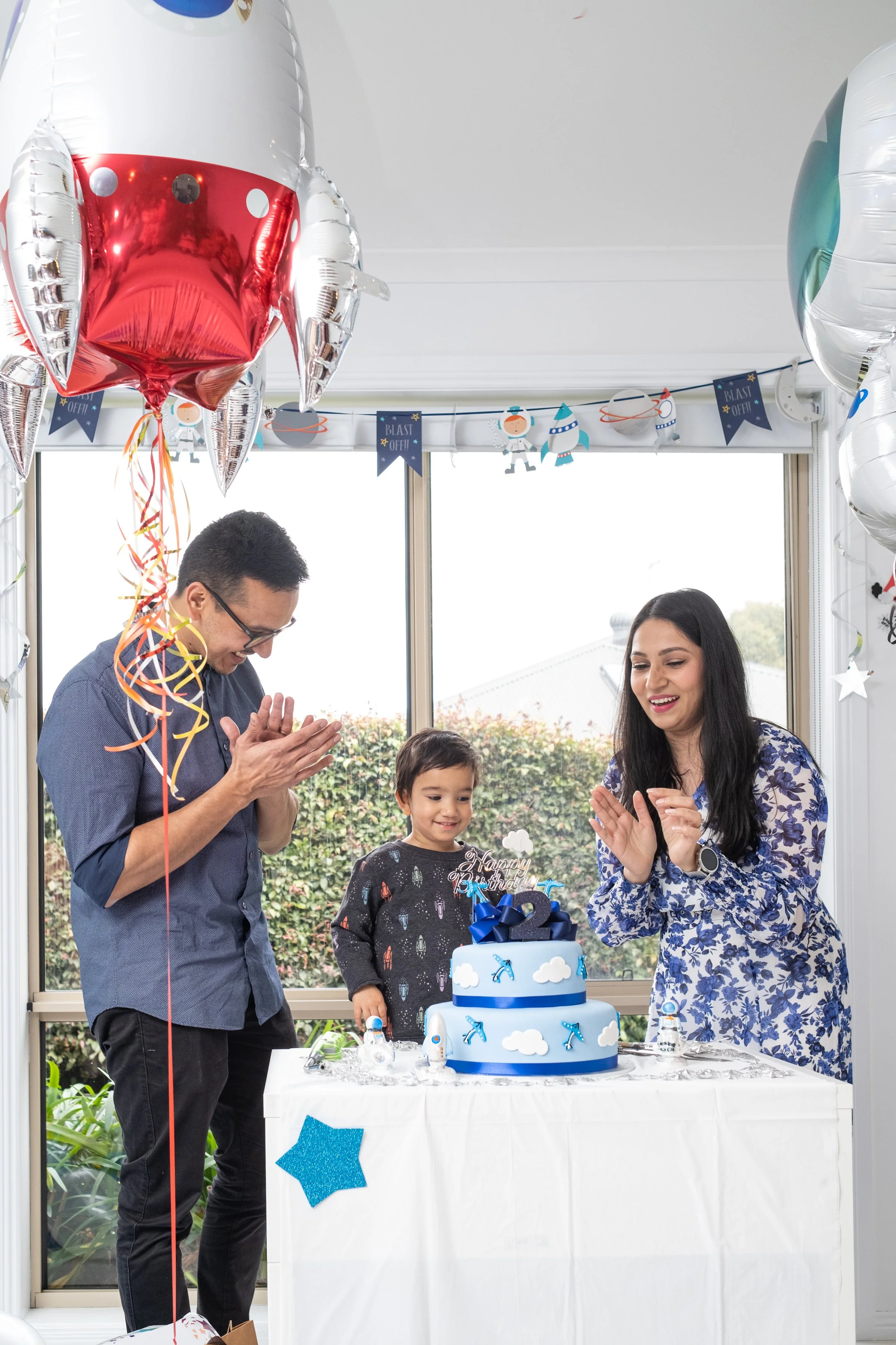 A family celebrating a birthday with a space-themed cake and decorations, featuring a boy and two adults clapping and smiling, in a bright room with large windows and space-themed banners.