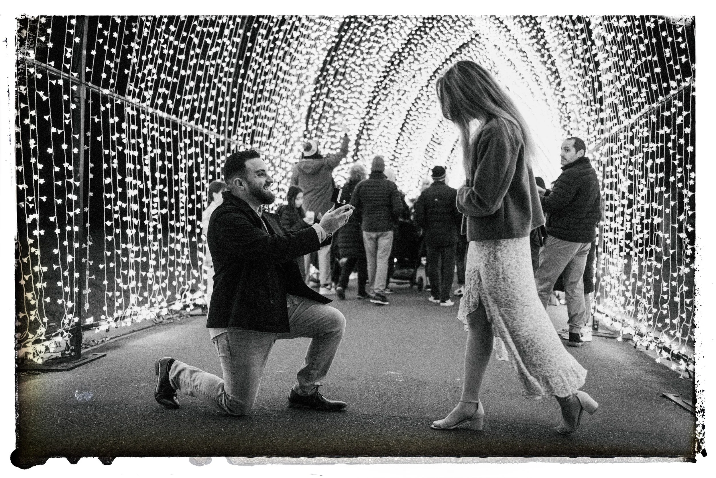 A man kneeling on one knee proposing to a woman in front of a tunnel of hanging lights, with other people walking in the background.