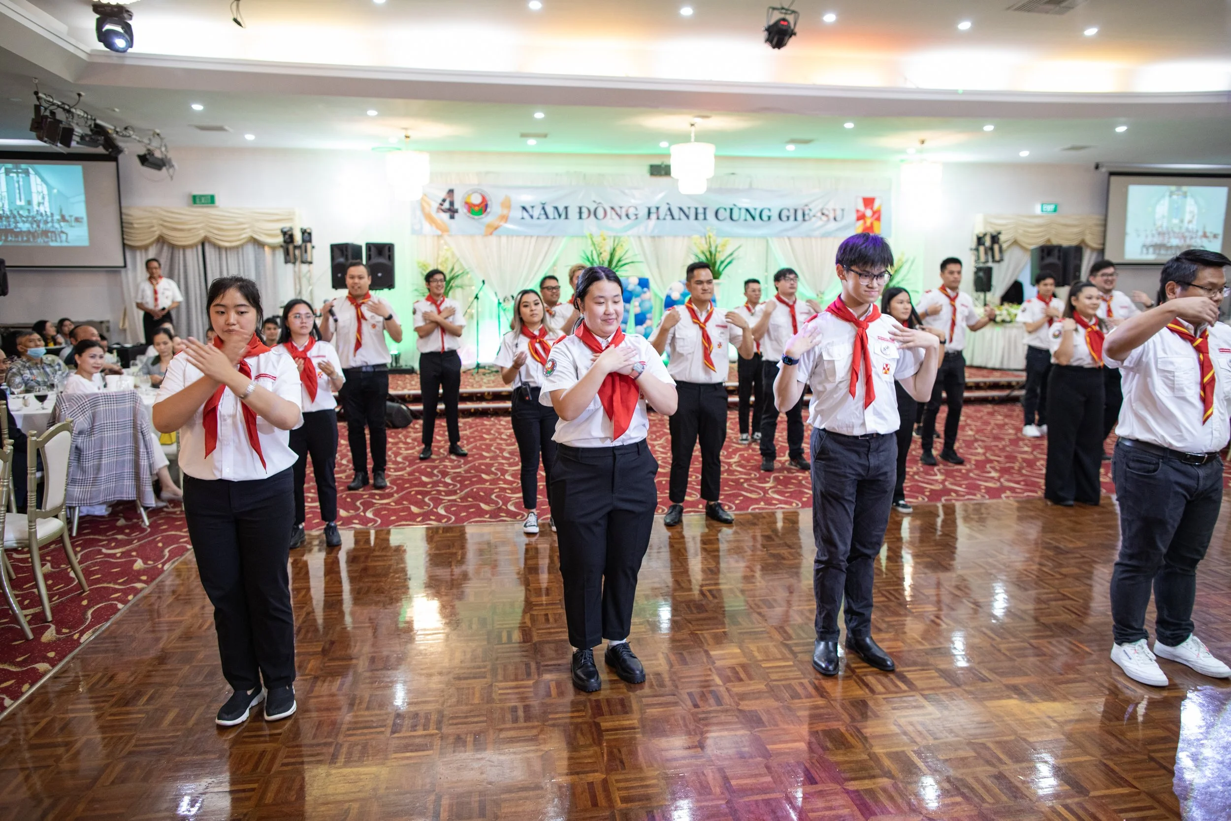 Group of people dressed in uniforms performing a dance or activity in a decorated banquet hall with a banner in the background.