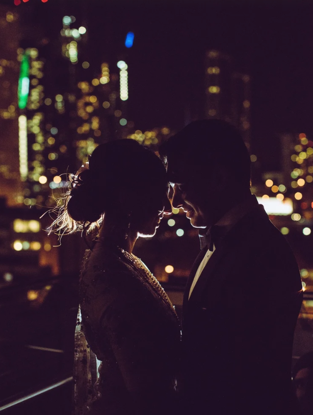 Silhouetted couple in wedding attire facing each other against a city skyline at night.
