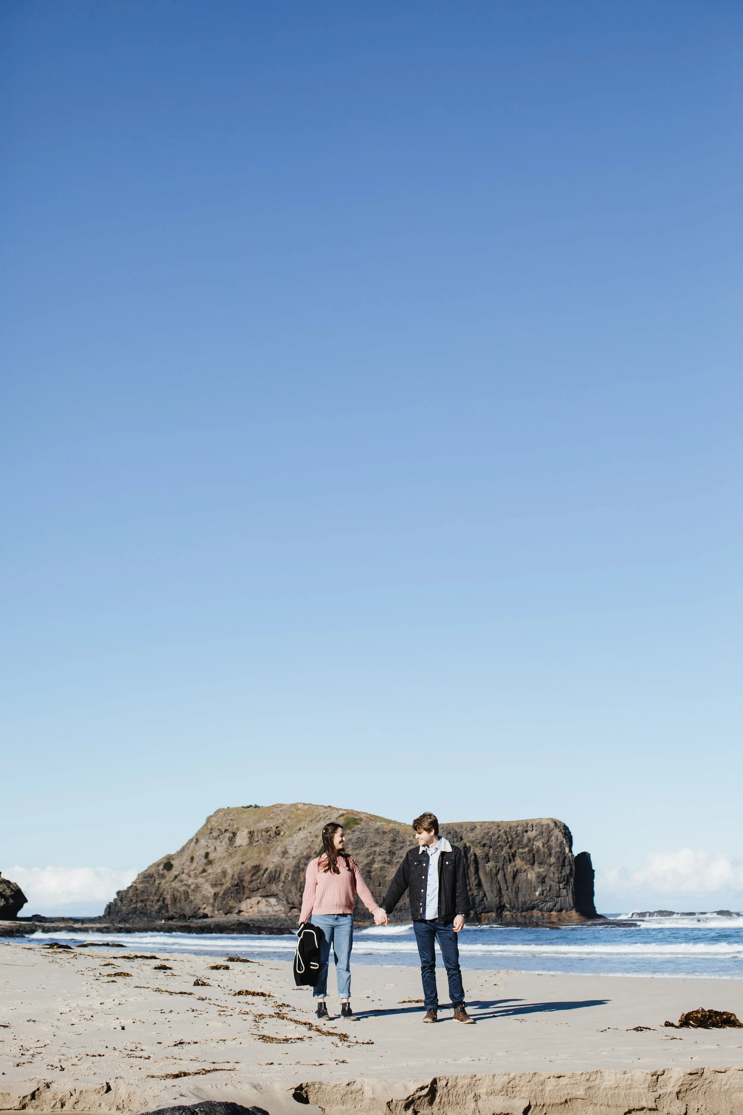 A young couple holding hands and walking along the beach with a rocky cliff in the background under a clear blue sky.