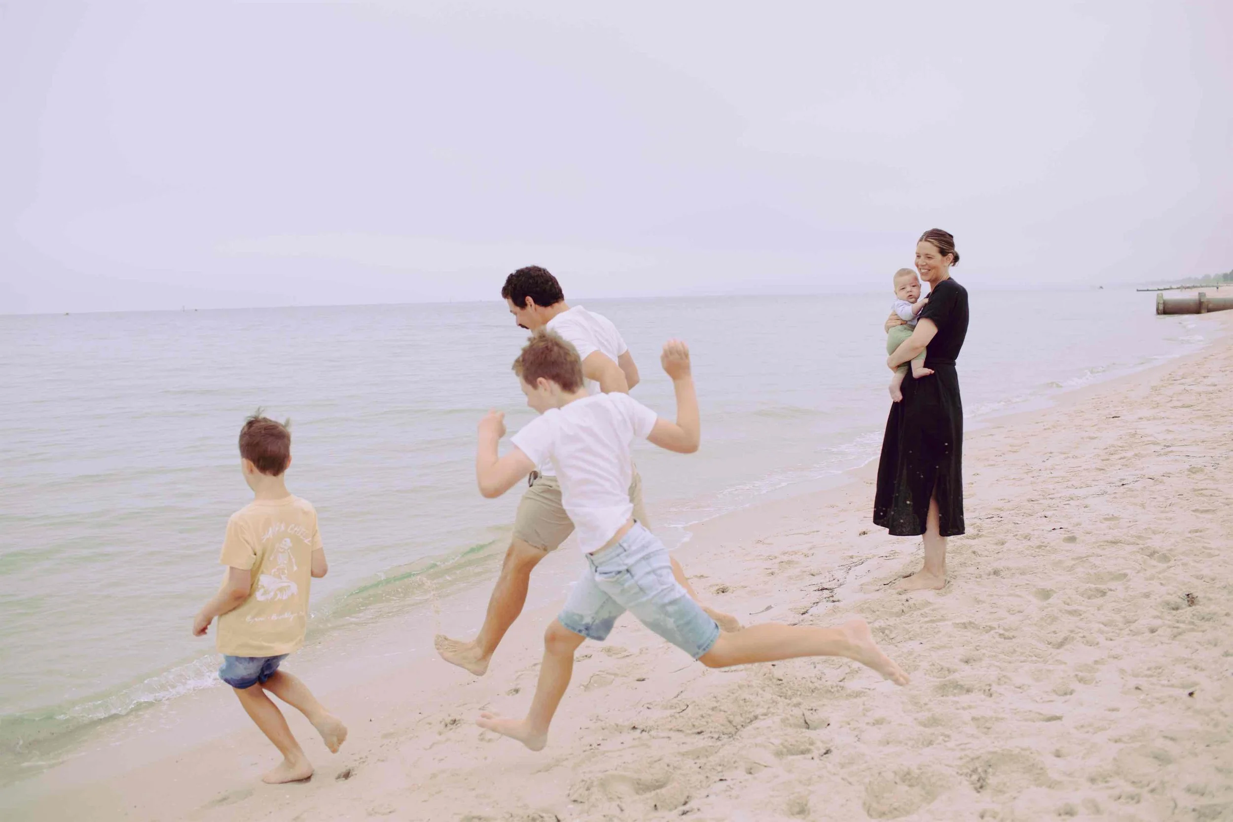 A group of children and a woman playing at the beach near the water, with the woman holding a baby and children jumping in the sand.