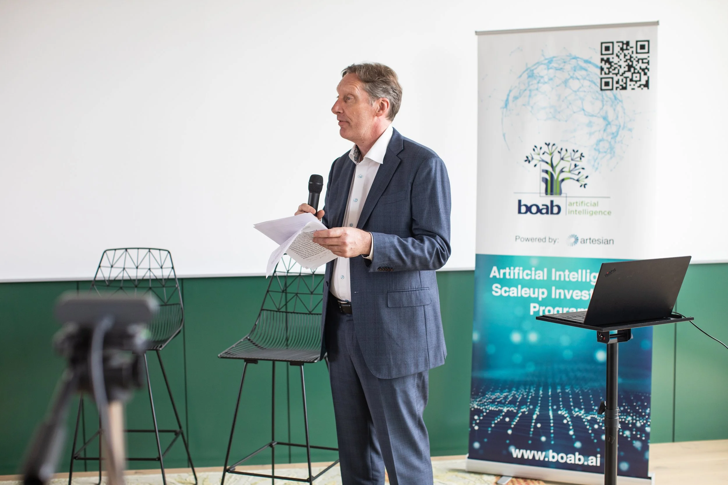 A man in a suit holding papers and a microphone, speaking at a conference or presentation with a banner about artificial intelligence and a laptop nearby.