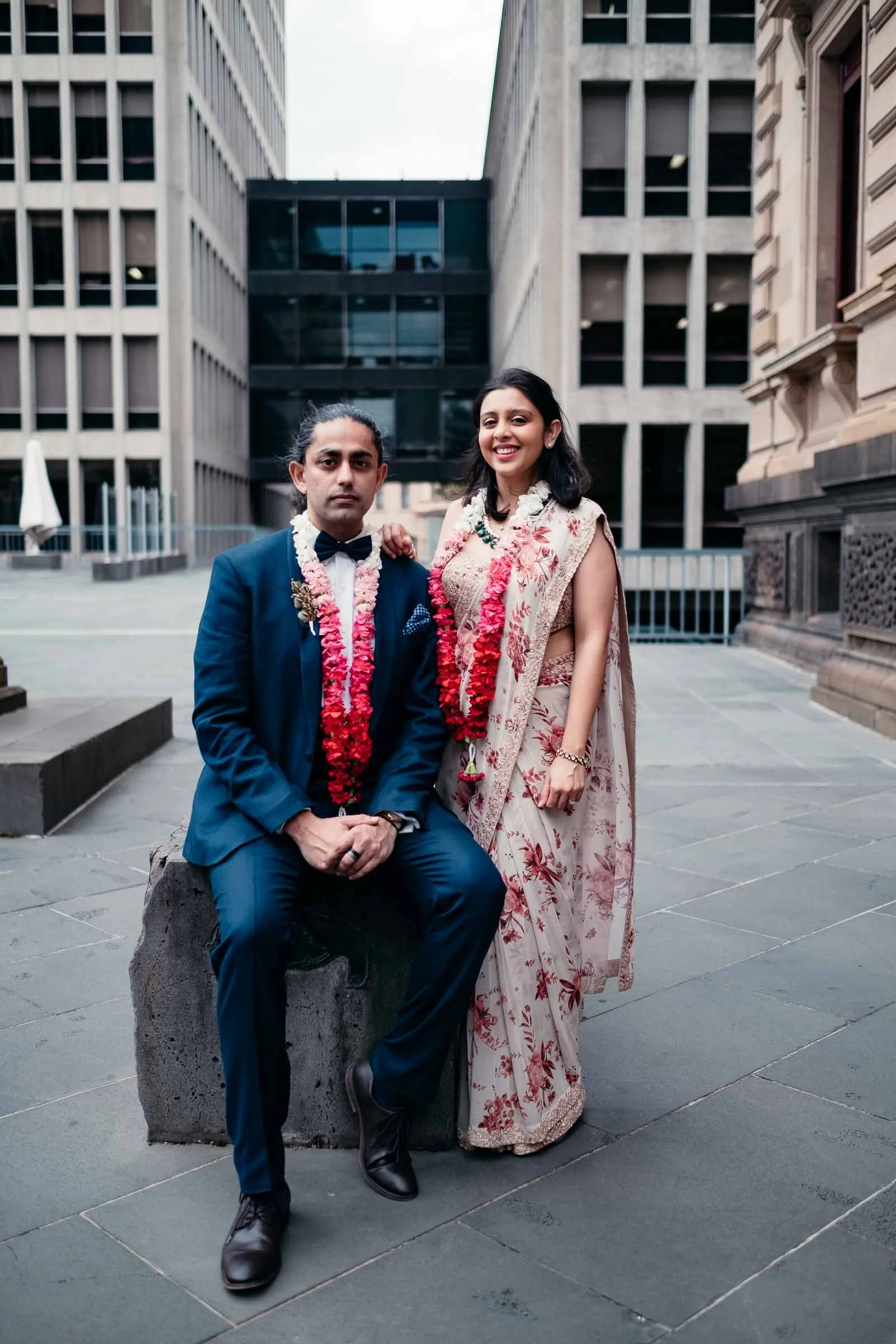 A couple dressed in traditional Indian attire. The man is wearing a blue suit with a bow tie, and the woman is in an elegant saree. Both are wearing flower garlands, standing outdoors in an urban setting with tall buildings in the background.
