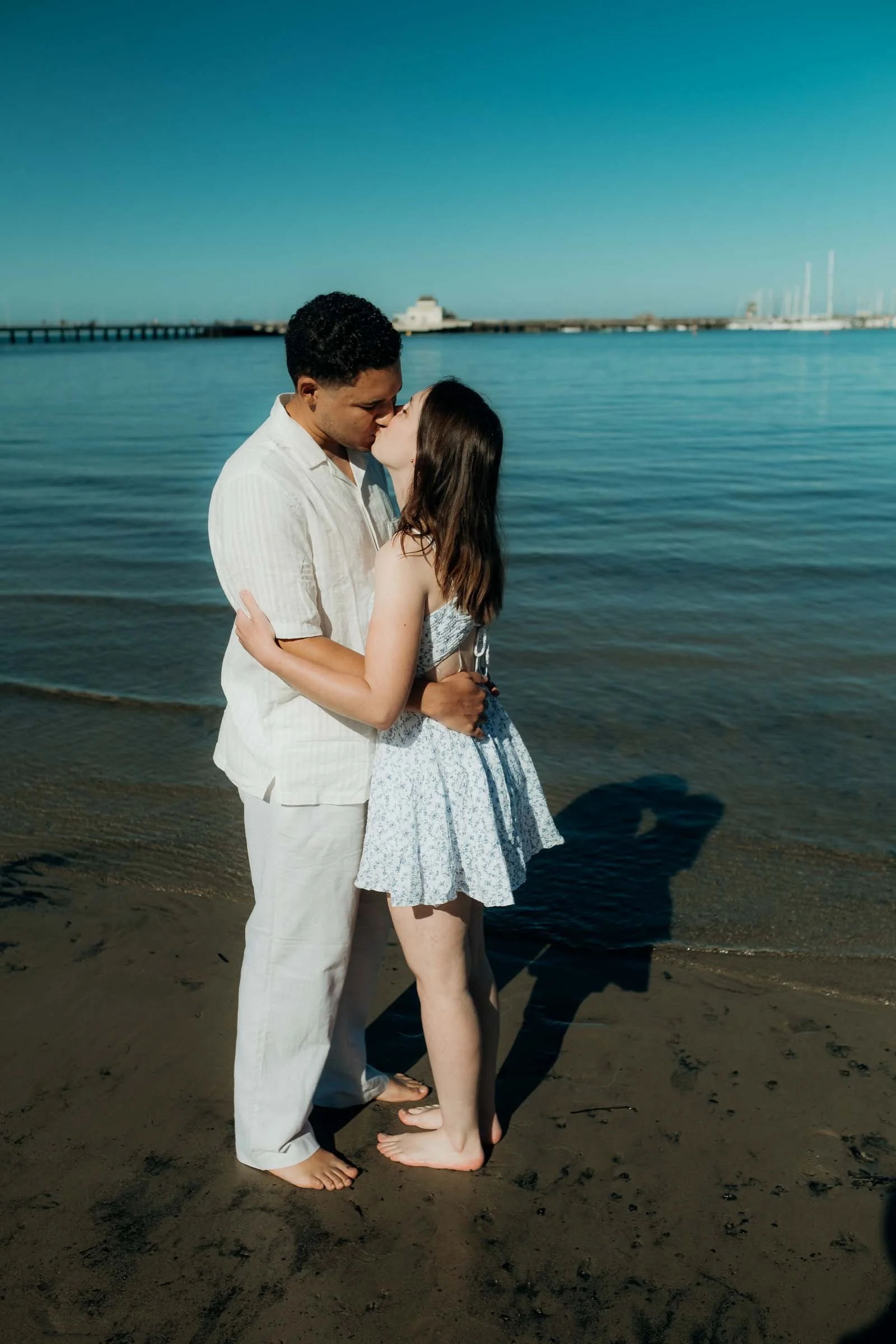 A couple standing barefoot on the beach, kissing by the water with a pier and sailboats in the background on a sunny day.