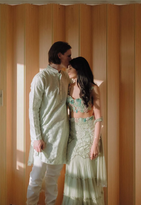 A couple dressed in traditional South Asian attire sharing an intimate moment with their foreheads touching in front of a wooden backdrop.