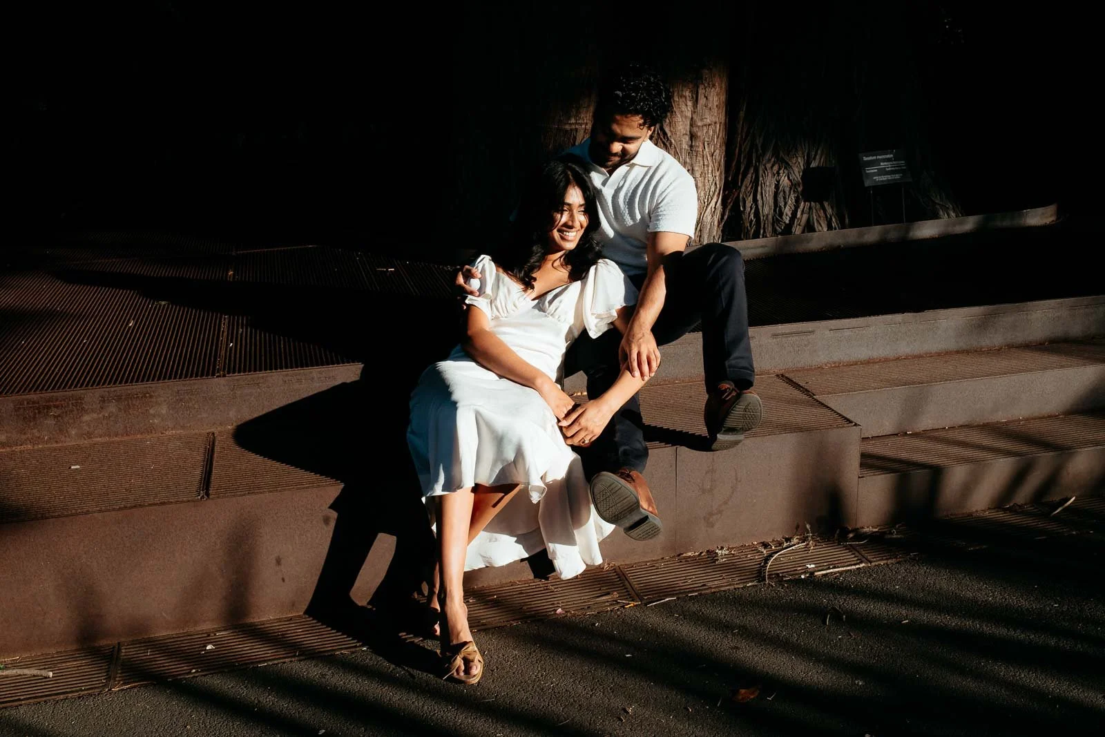 A smiling couple sitting on a park bench, with the woman dressed in a white dress and the man in a white shirt and dark pants, sharing an intimate moment during sunset.