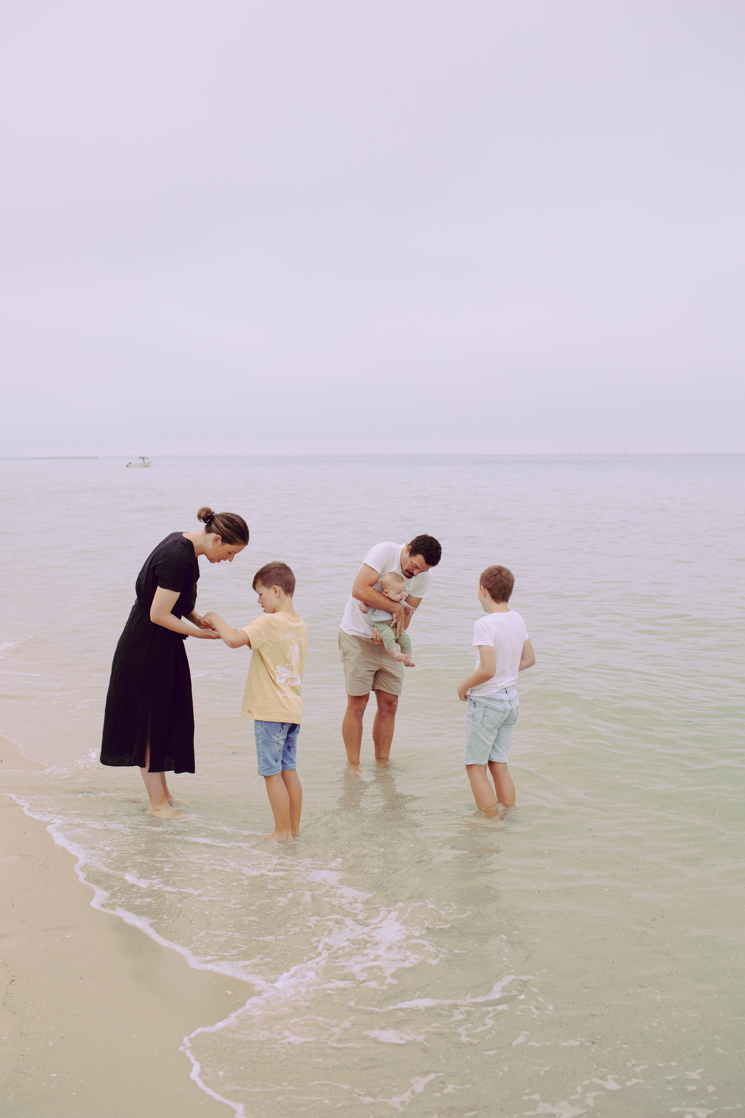 A woman, man, and children standing in shallow water at the beach, holding hands and looking down, with overcast sky and distant boats on the horizon.