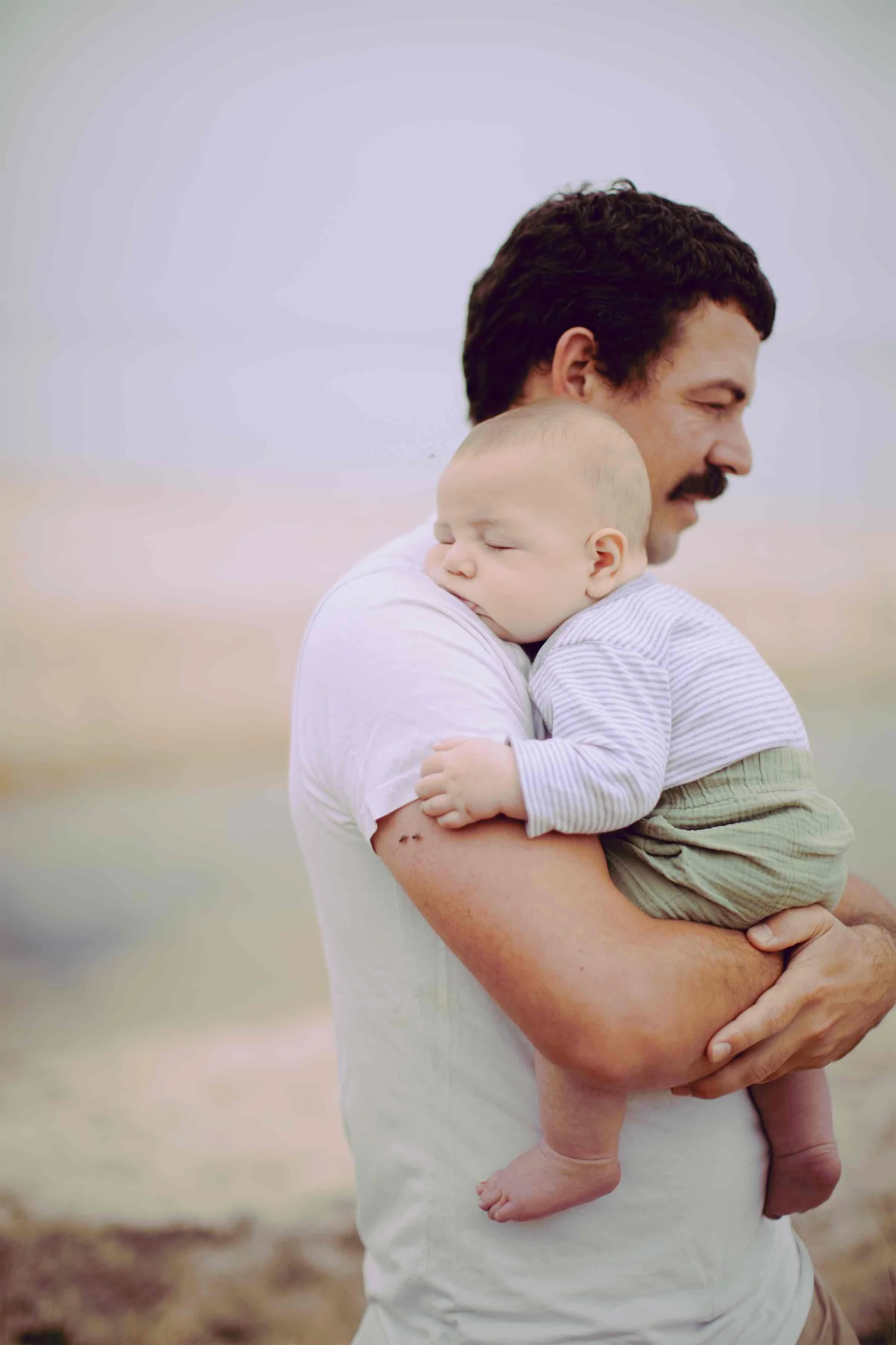 A man holding a sleeping baby outdoors, both smiling and wearing casual white and beige clothes.