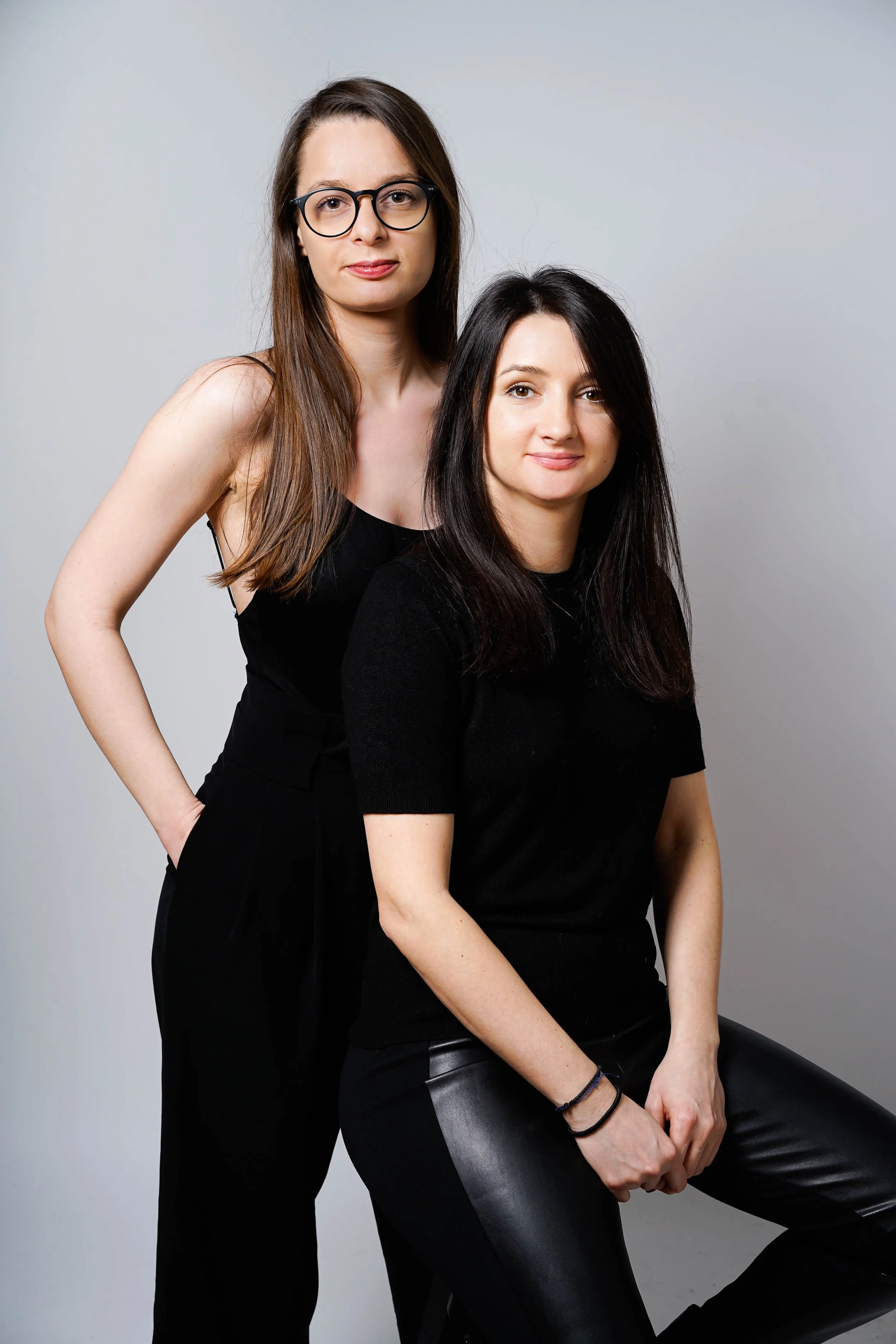 Two women, one seated and one standing behind her, both dressed in black, against a plain gray background.