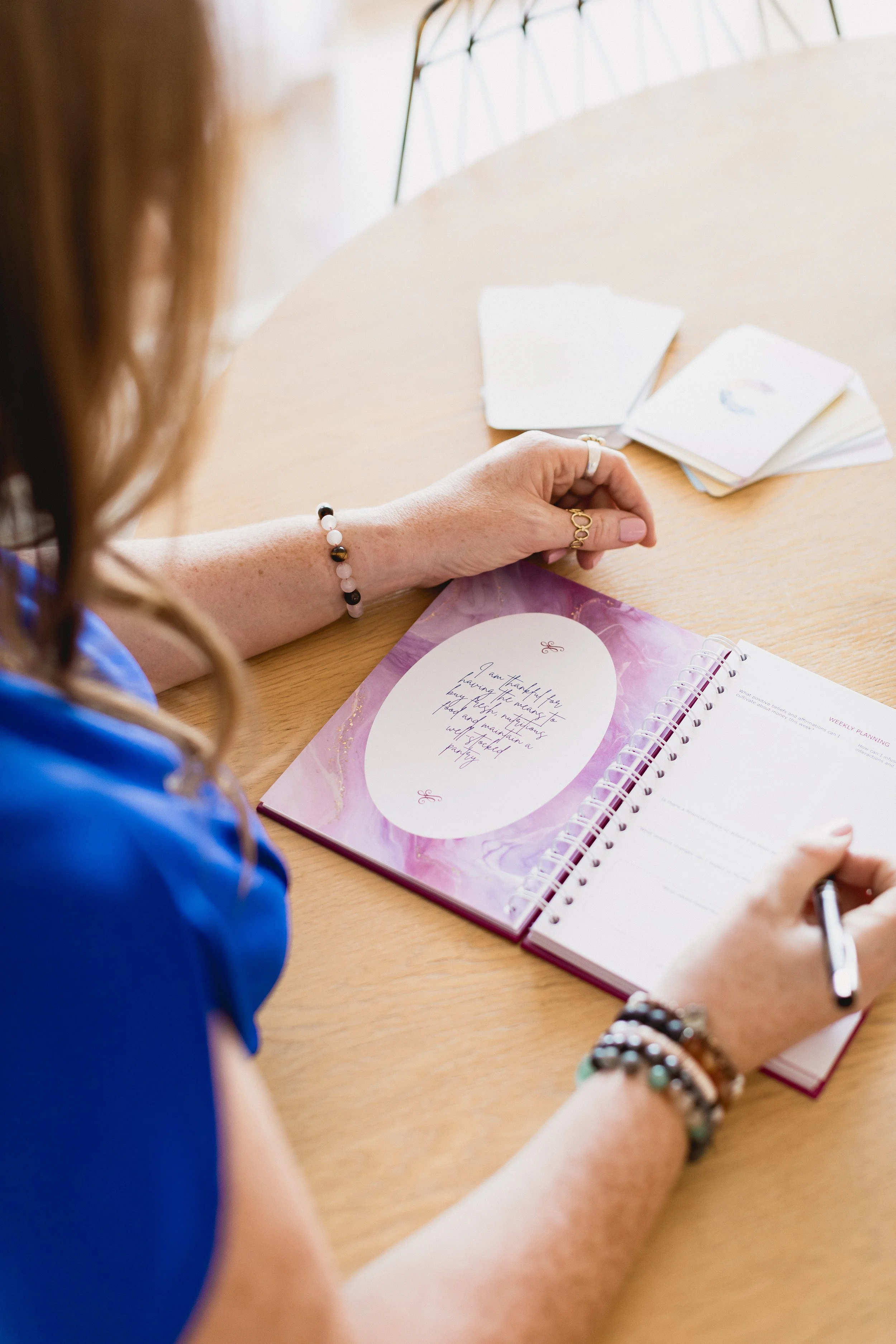 A person with bracelets on their wrist writing in a spiral planner on a wooden table, with a stack of blank and patterned cards nearby.