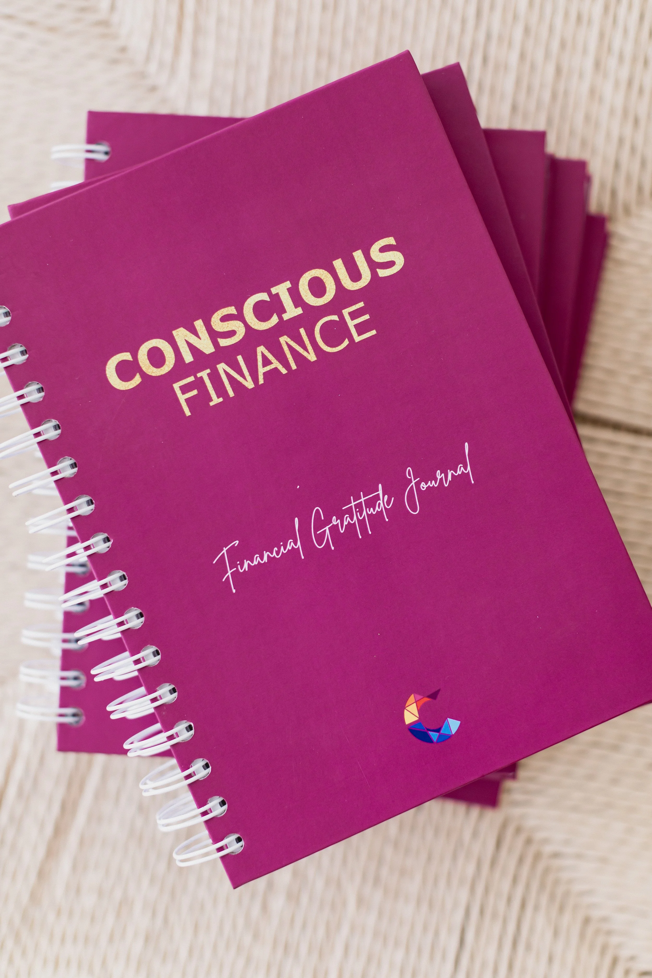 Close-up of a pink spiral-bound notebook titled 'Conscious Finance' with the subtitle 'Financial Gratitude Journal' and a colorful geometric logo at the bottom, placed on a light-colored woven surface.