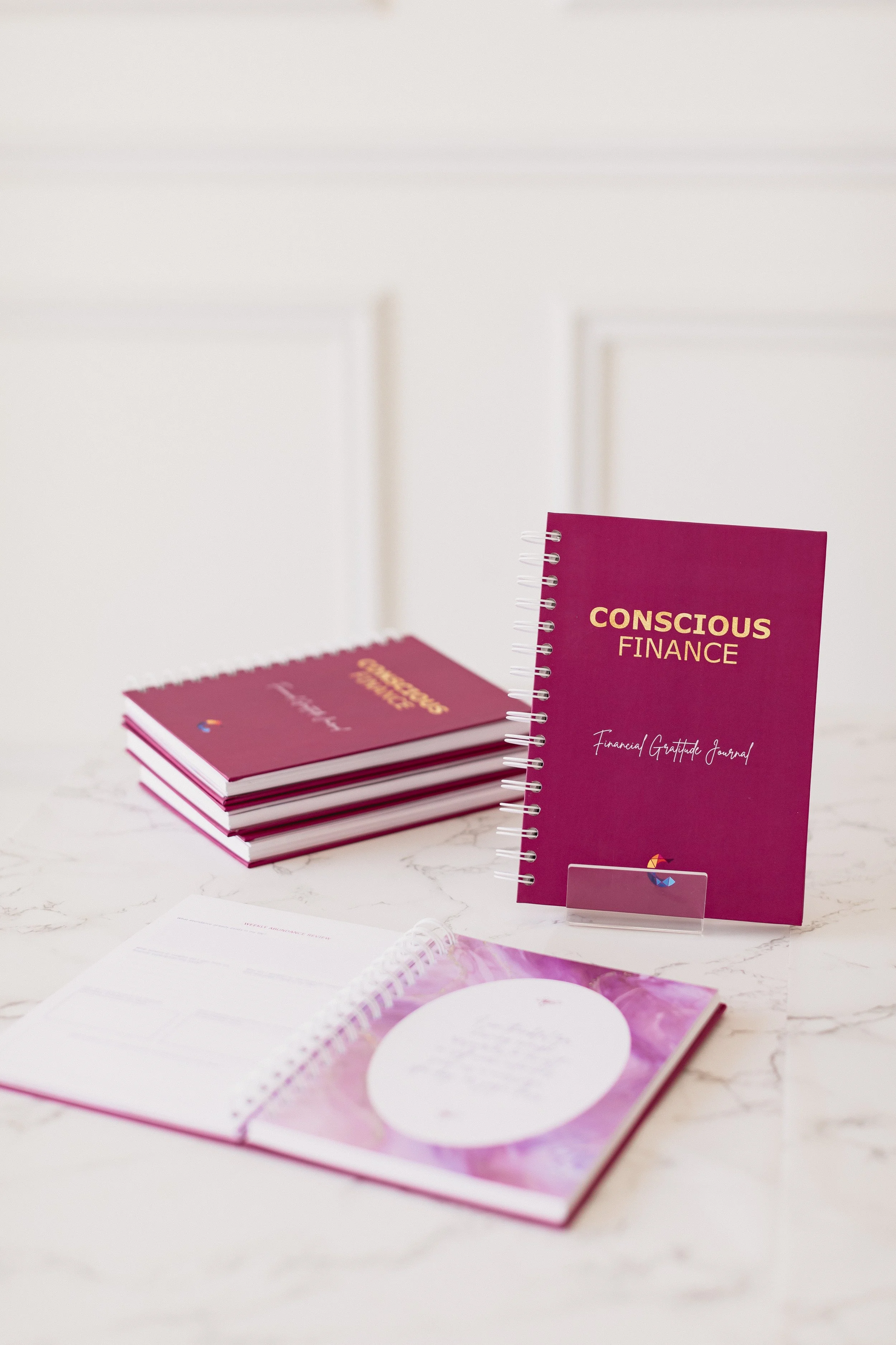 A table displaying pink notebooks titled "Conscious Finance" and "Financial Gratitude Journal," along with open planners and a clear sign with a rainbow-colored pie chart logo.