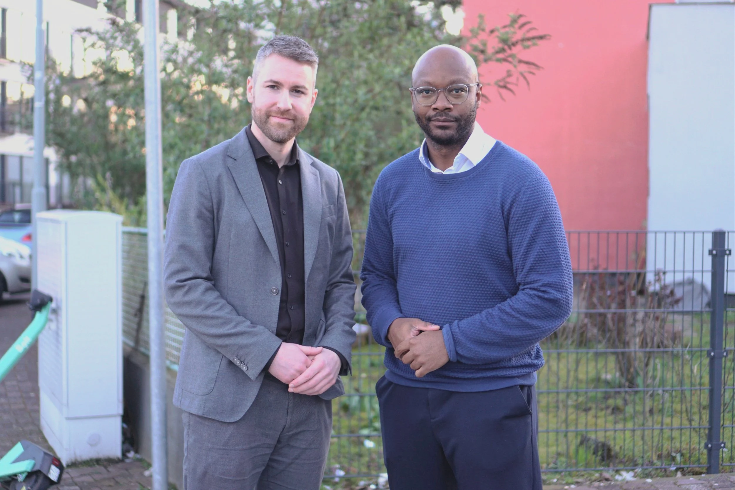Zwei Männer stehen nebeneinander im Freien, einer in einem grauen Anzug und der andere in einem blauen Pullover. Im Hintergrund ist ein Gartenzaun und ein Gebäude zu sehen.
