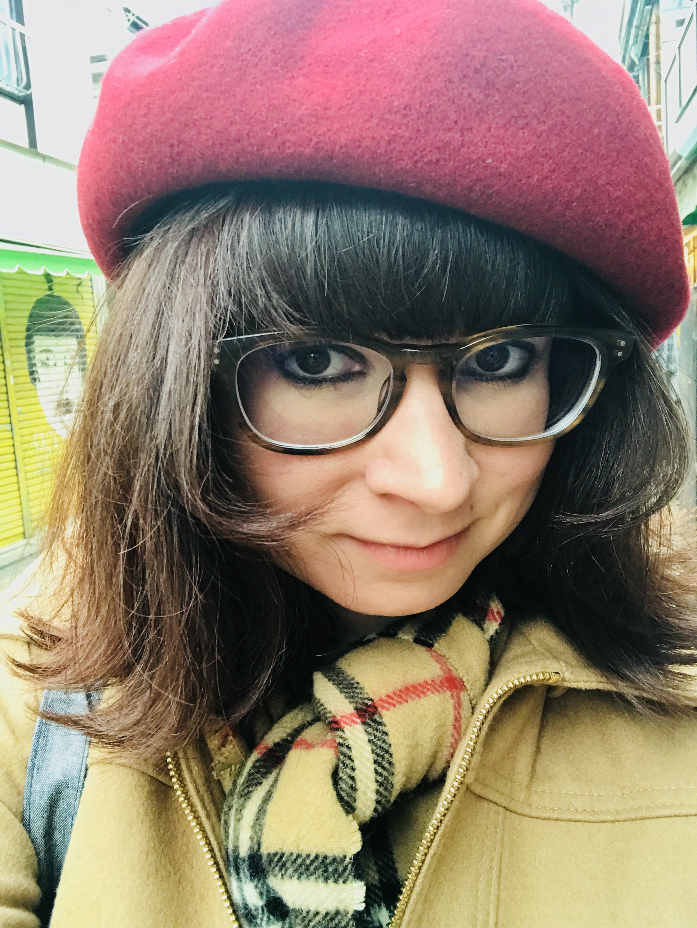Close-up of a woman with dark hair, wearing glasses, a red beret, a beige jacket, and a plaid scarf, outdoors in a city environment.