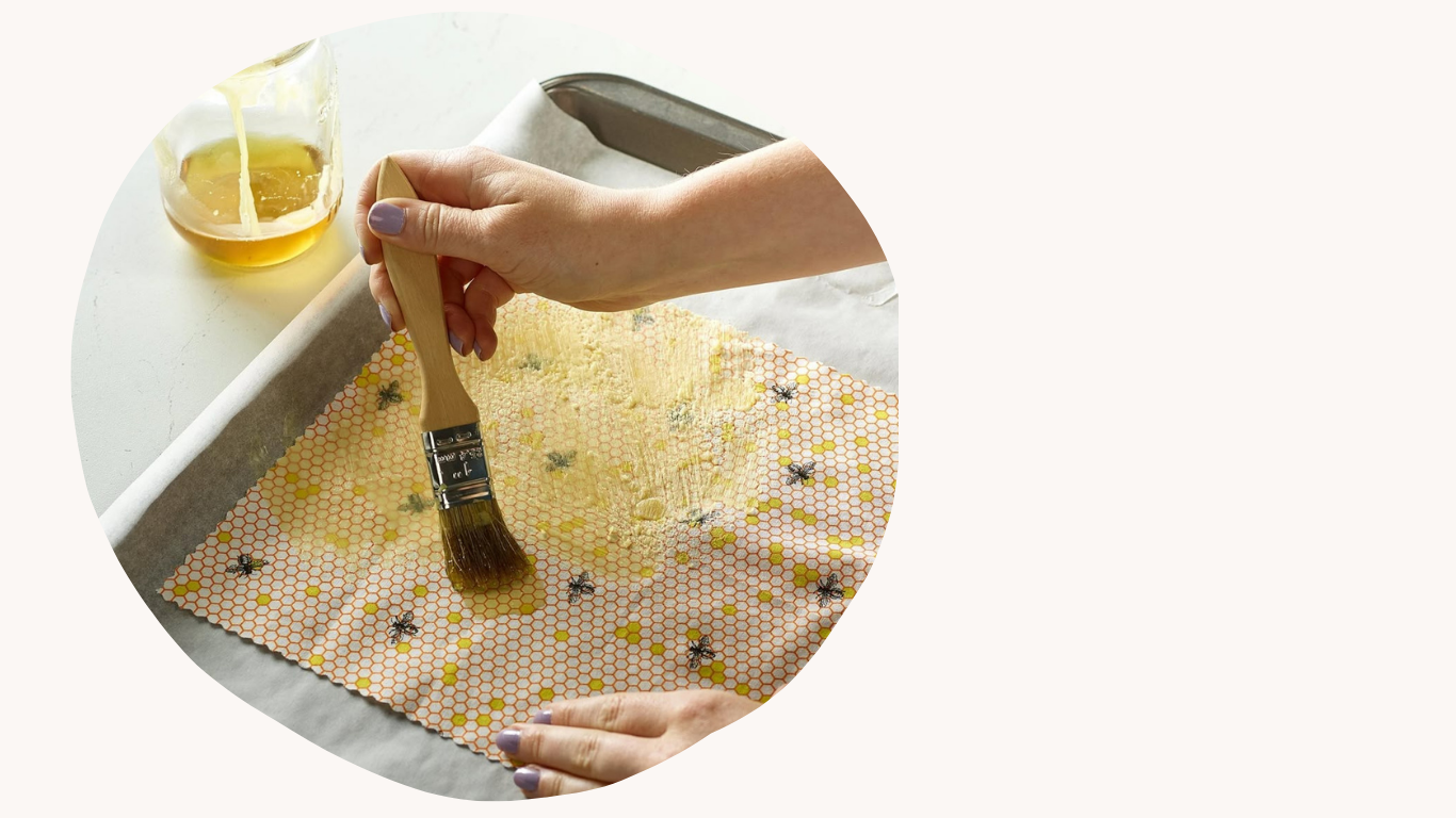 Person using a brush to apply beeswax wrap with a honeycomb and bee pattern to a sheet of parchment paper, with a glass of honey and a butter knife nearby.