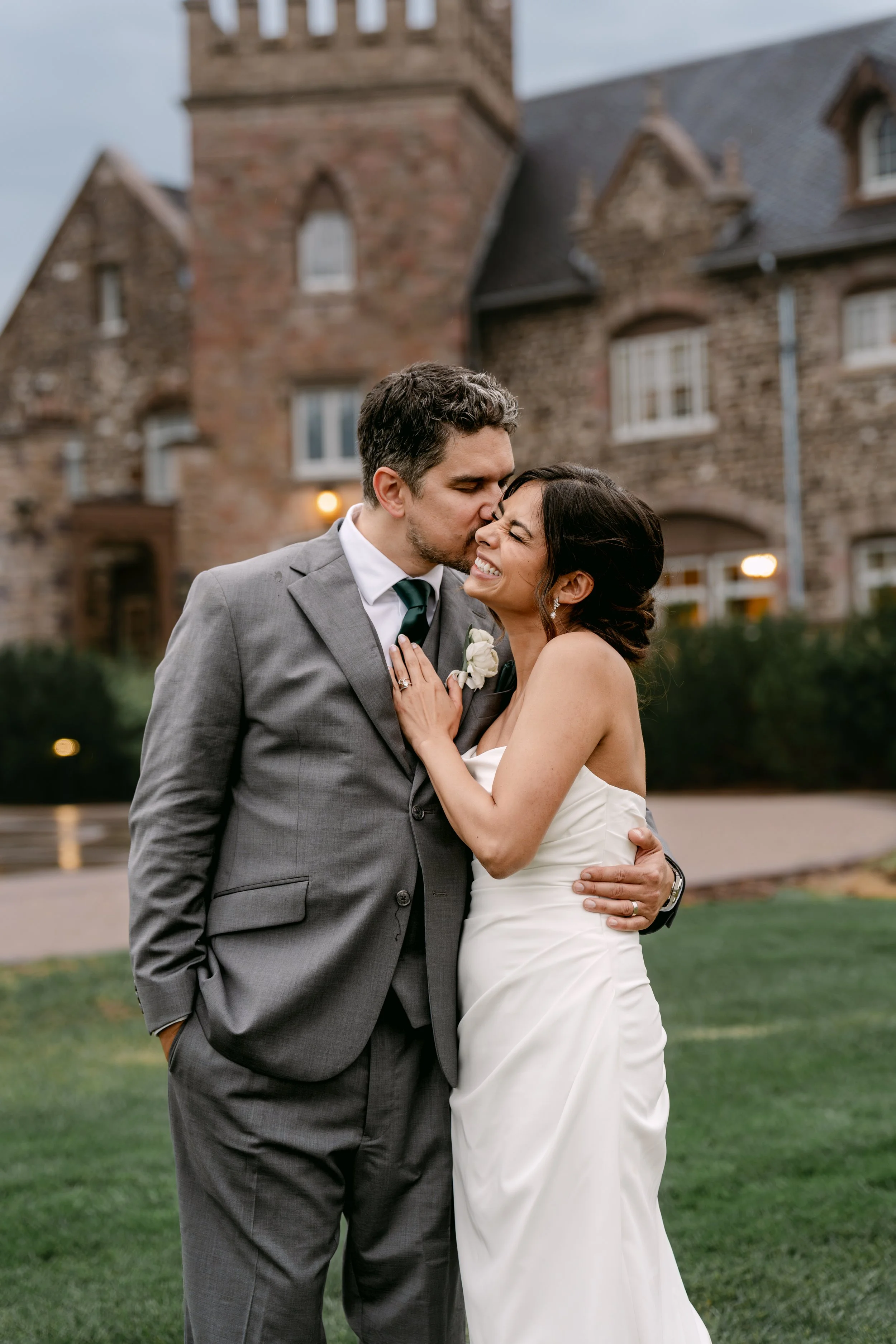 A smiling bride embraces her groom as he kisses her cheek, with a rustic stone building in the background.