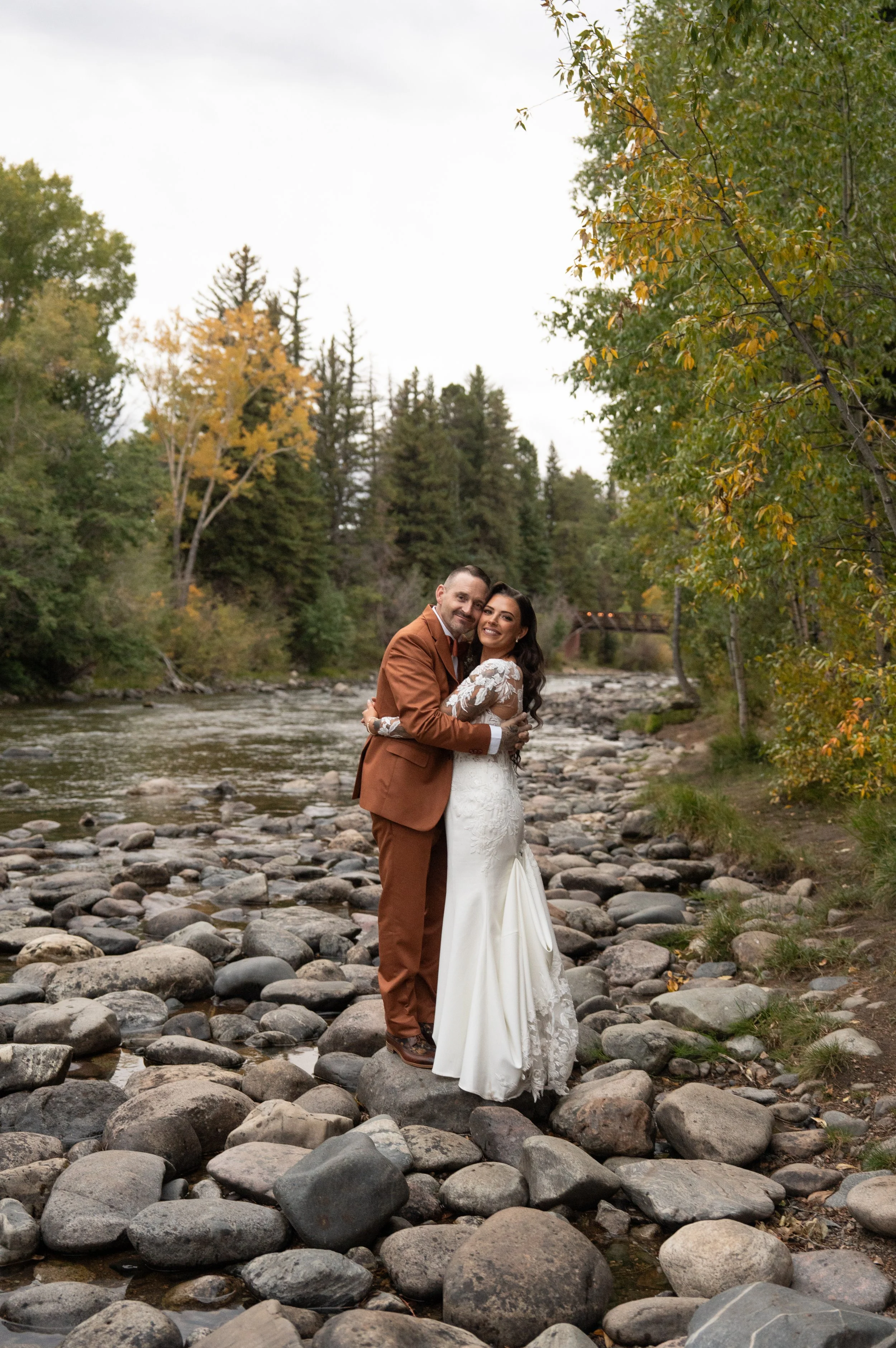 A bride and groom embrace on a rocky riverbank surrounded by lush trees showing early signs of autumn. The bride wears a long-sleeved lace wedding gown with a fitted silhouette, while the groom wears a rust-colored suit.