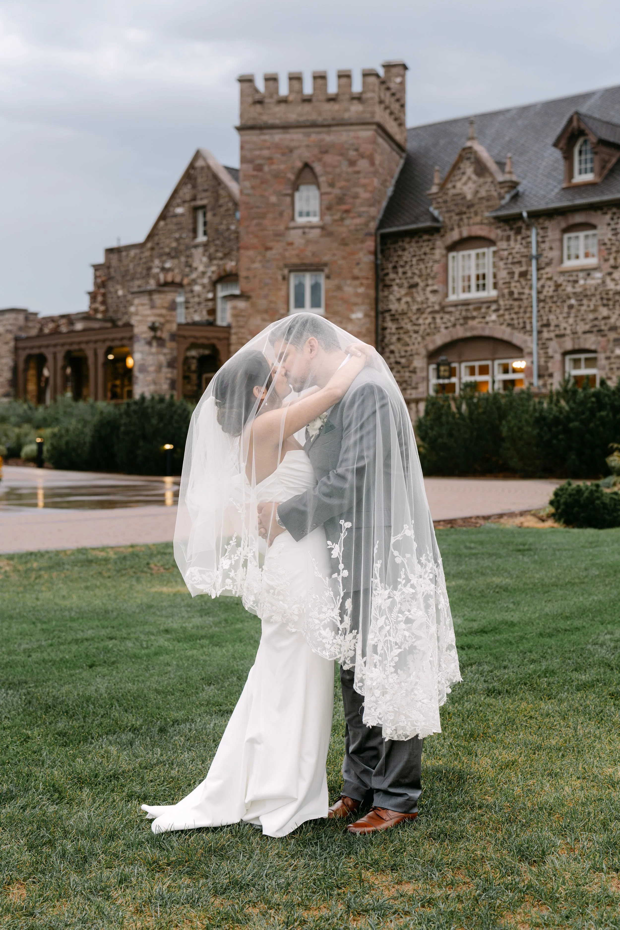 A bride and groom kiss beneath her veil on a lawn in front of a rustic stone venue.