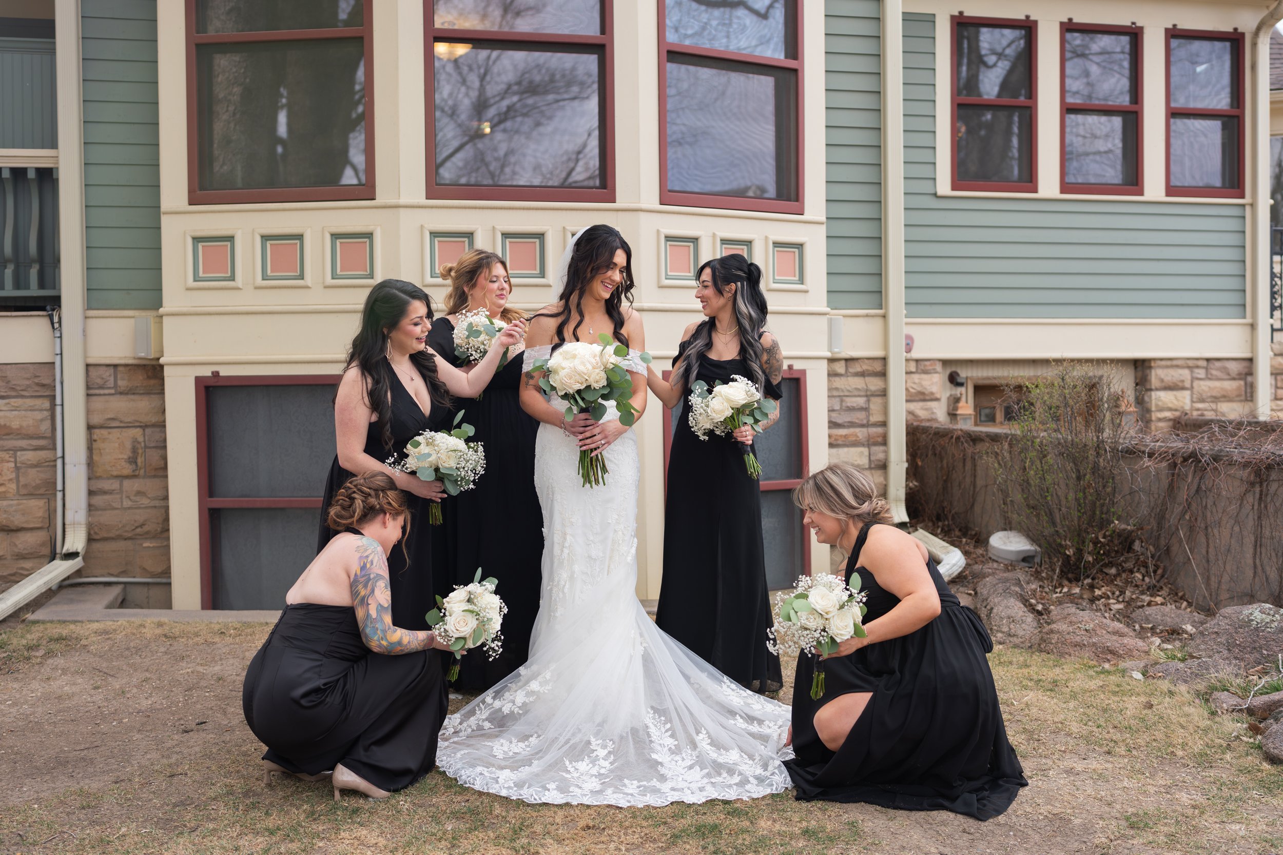 A wedding party celebrating outdoors, with the bride and groom at the center, surrounded by bridesmaids and groomsmen. The bride is laughing and holding a bouquet, and the groom is smiling at her. Bridesmaids are in pink dresses and holding bouquets, while groomsmen are in blue suits with pink bow ties.
