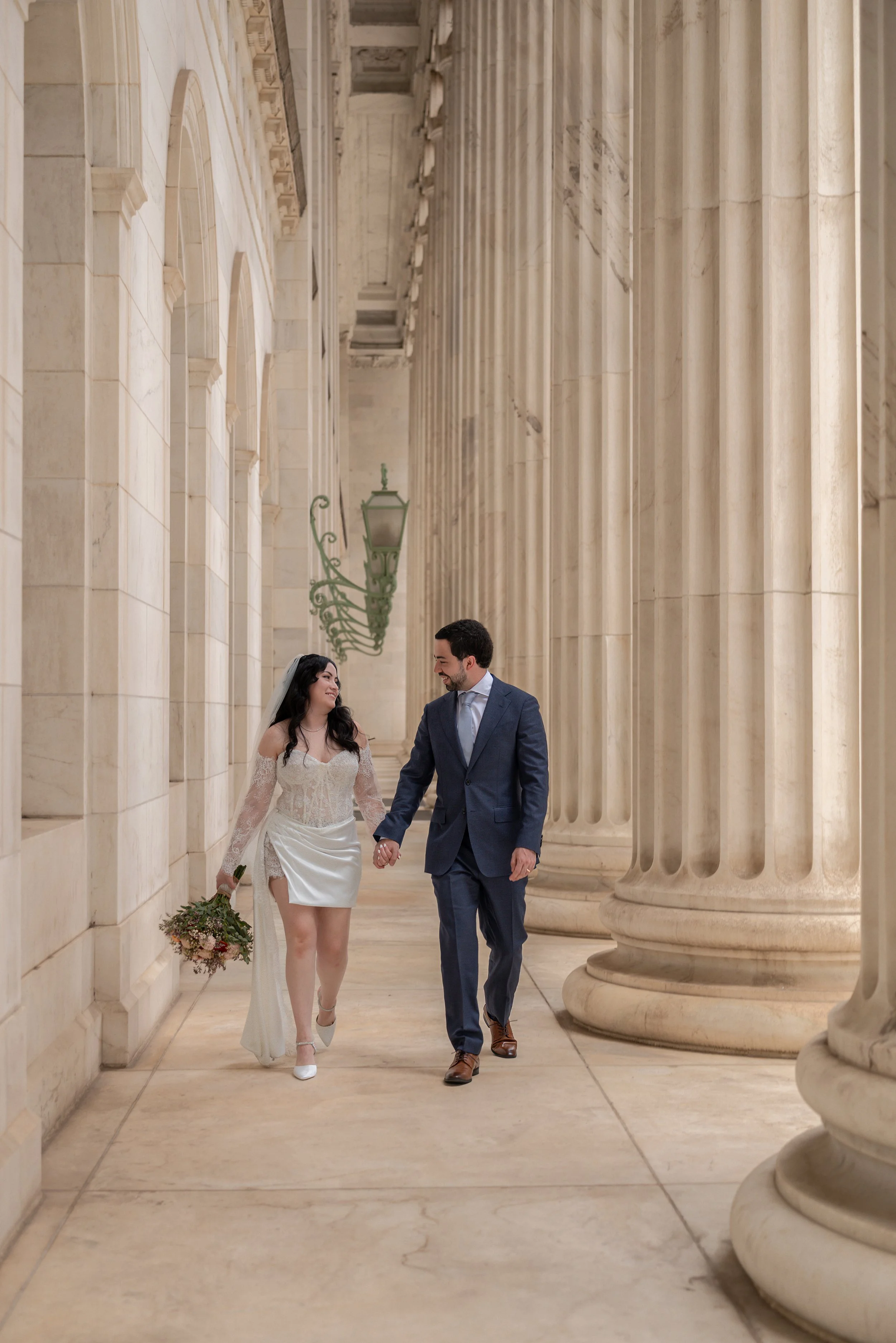 A bride and groom walk hand in hand down a grand marble corridor lined with tall columns and elegant arches. The bride, wearing a short white dress with lace sleeves, a flowing veil, and heels, holds her bouquet by her side. 