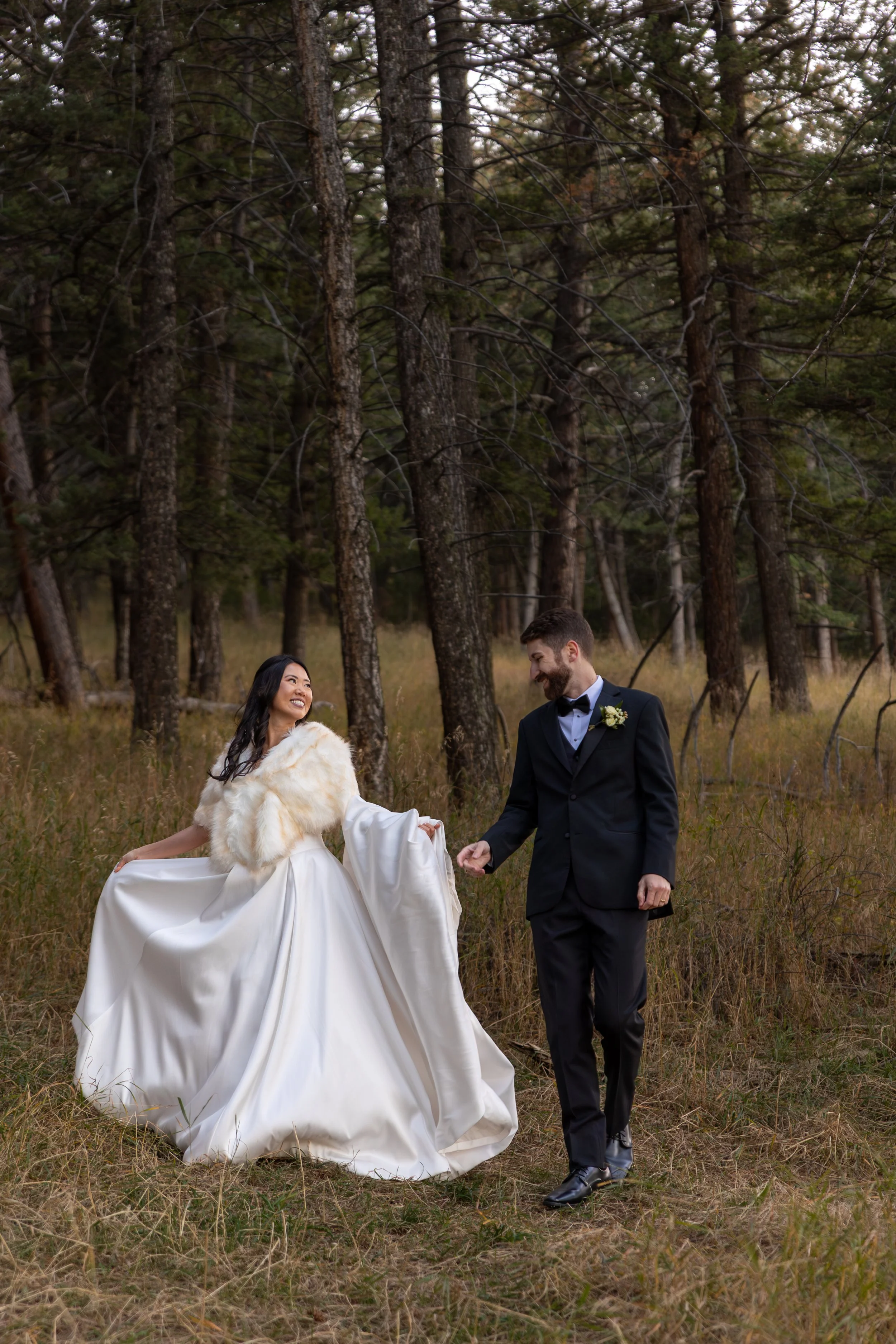A bride and groom walk hand in hand through a forest clearing, surrounded by tall pine trees and golden grass. The bride wears a flowing satin gown with a soft fur shawl draped over her shoulders, smiling joyfully as she lifts her dress slightly.