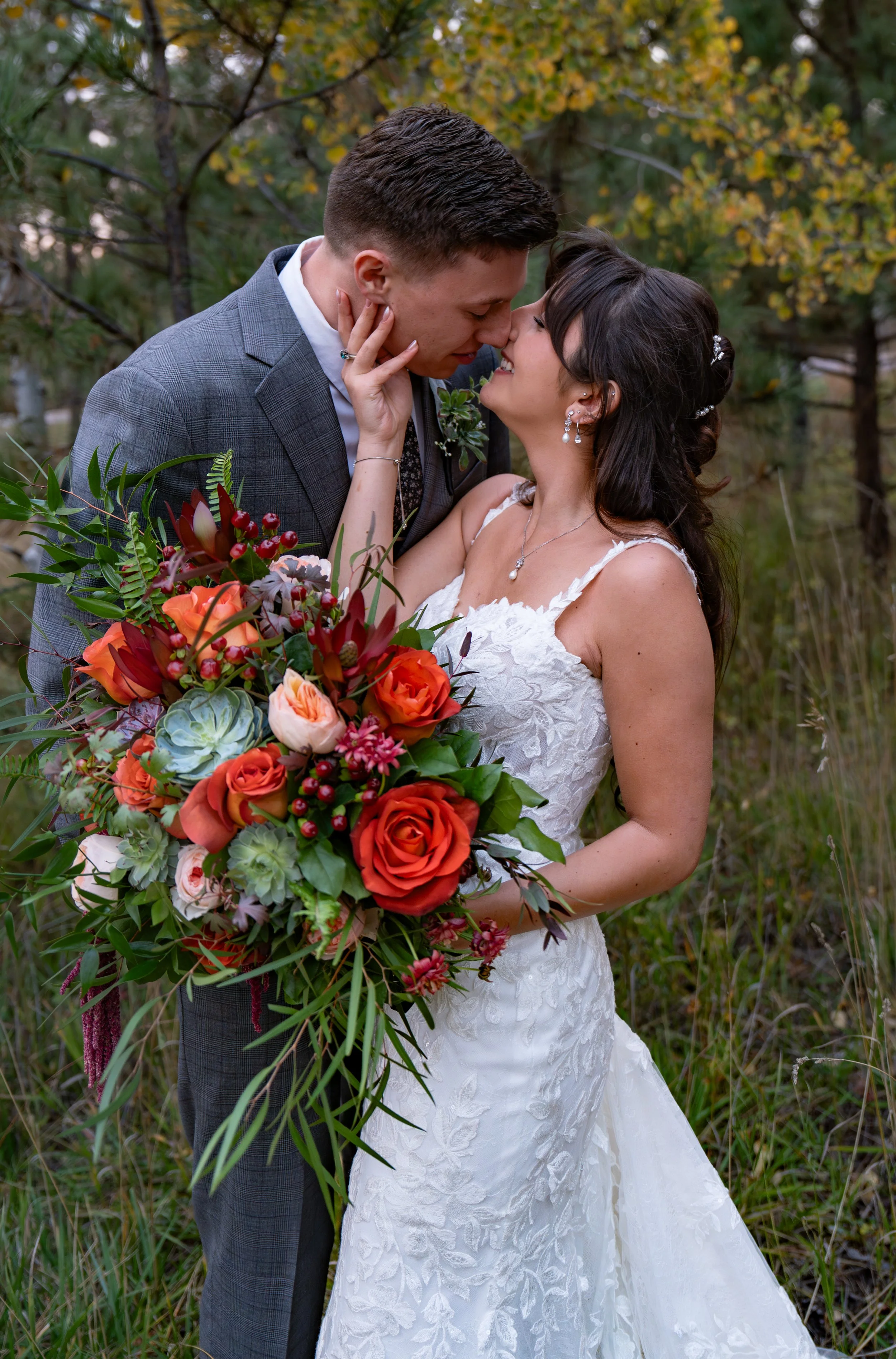 A bride and groom share a tender moment in a forest setting, standing close as they lean in for a kiss. The bride, in a lace wedding gown with delicate straps, holds a vibrant bouquet filled with red, orange, and blush roses, greenery, and succulents