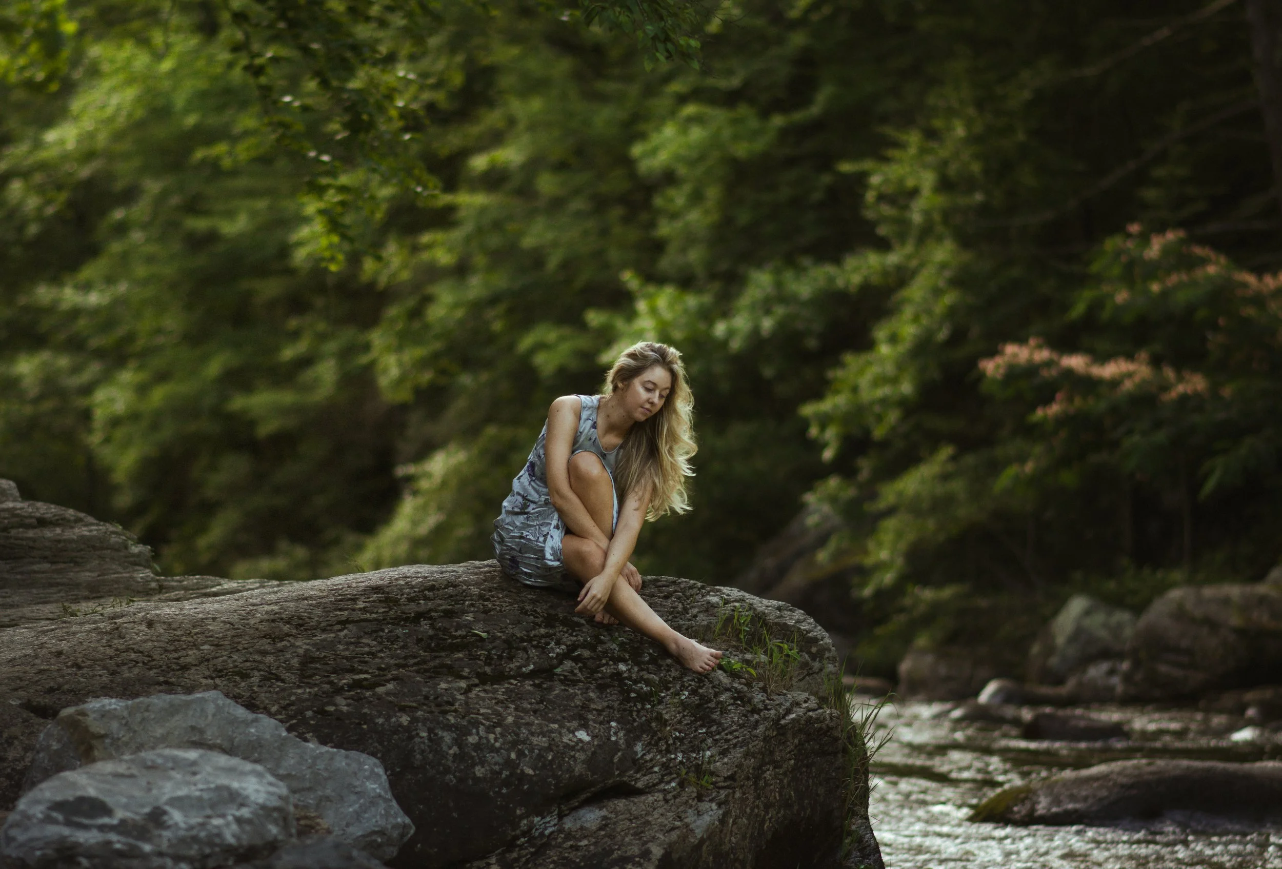 A young woman with long blonde hair, wearing a sleeveless dress, sitting on a fallen tree trunk by a river in a lush green forest, looking down at the water.