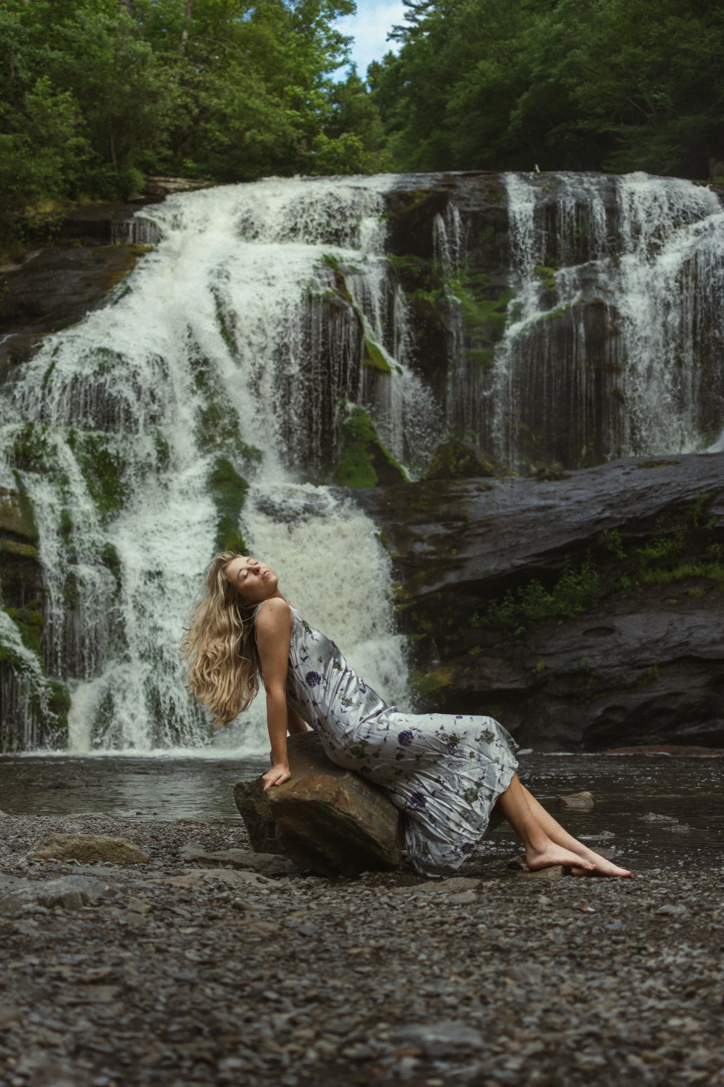 A woman with long blonde hair, wearing a white dress with purple flowers, sitting on a large rock in a stream with a waterfall and green forest in the background.