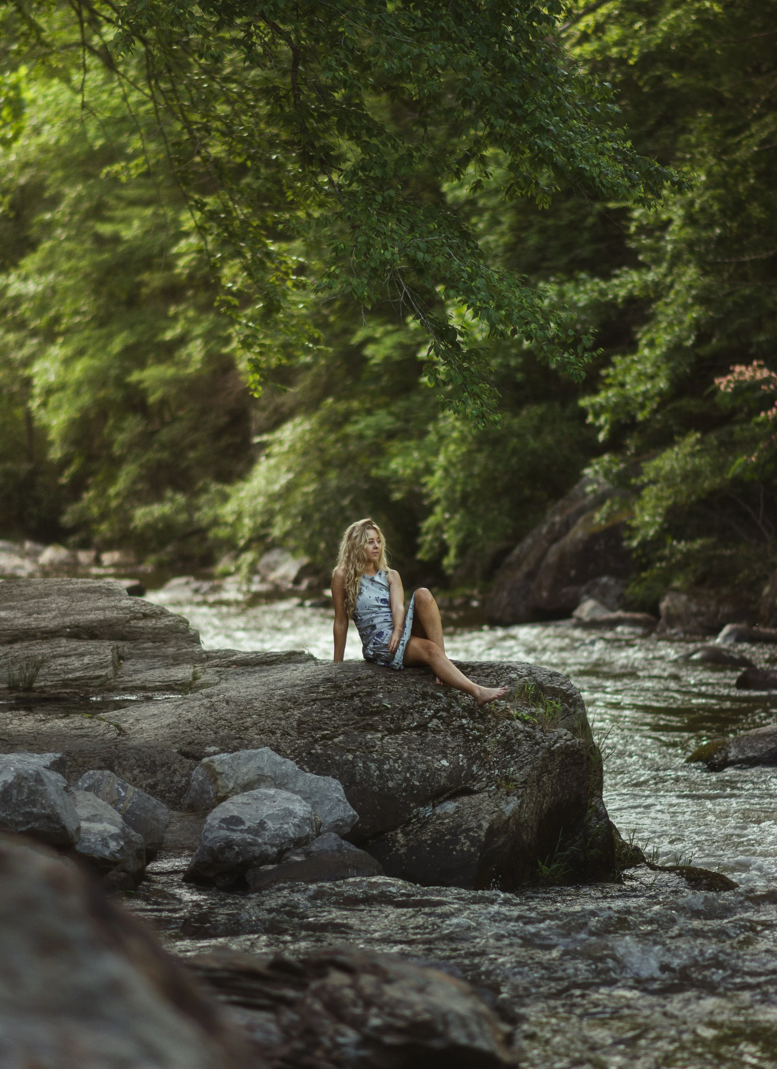 A woman with long blonde hair sitting on a large rock in a creek surrounded by green trees and foliage.