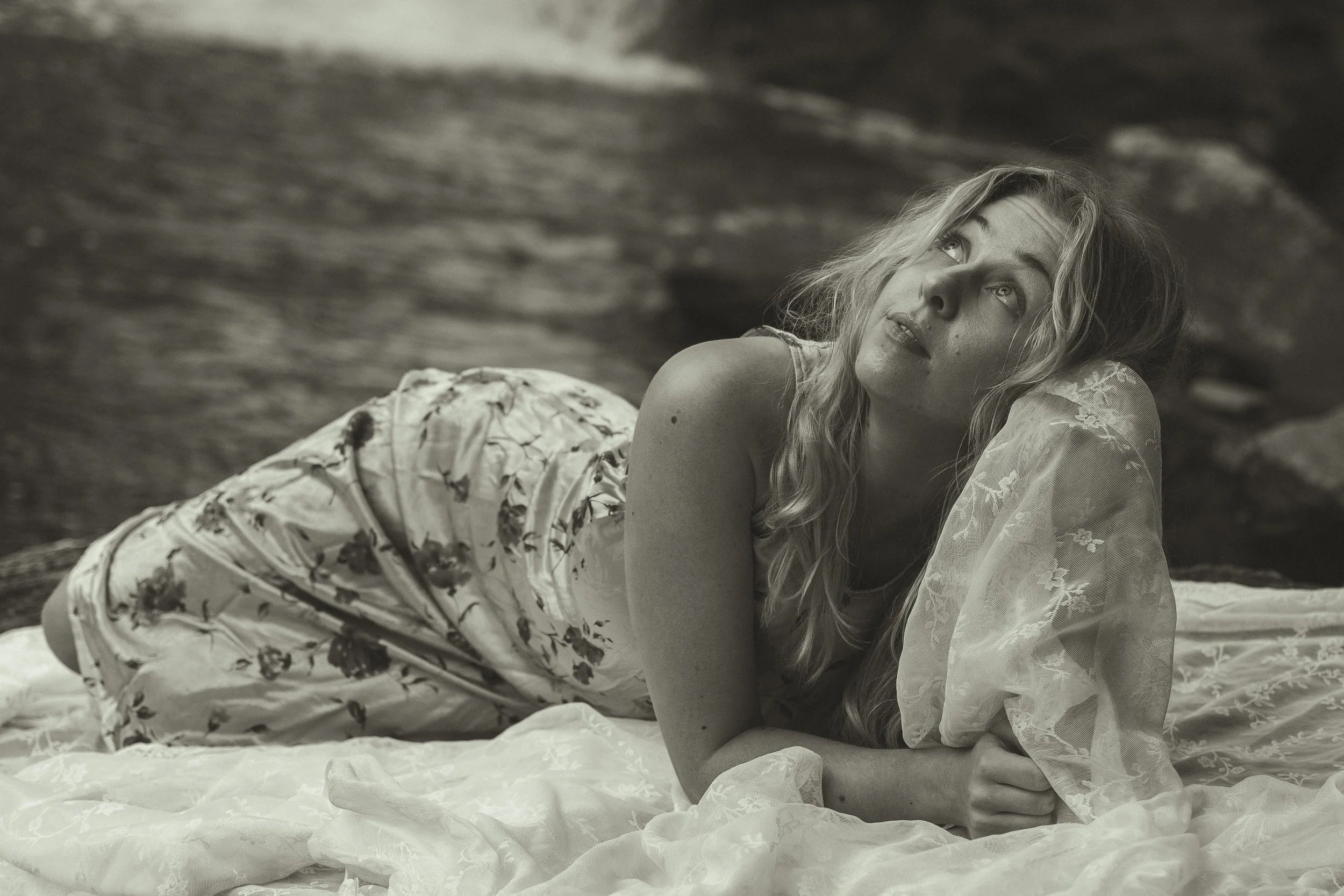 A woman with wavy blonde hair lying on her side on a bed, gazing upwards, wearing a sheer lace top and patterned pants, in a black and white photograph.