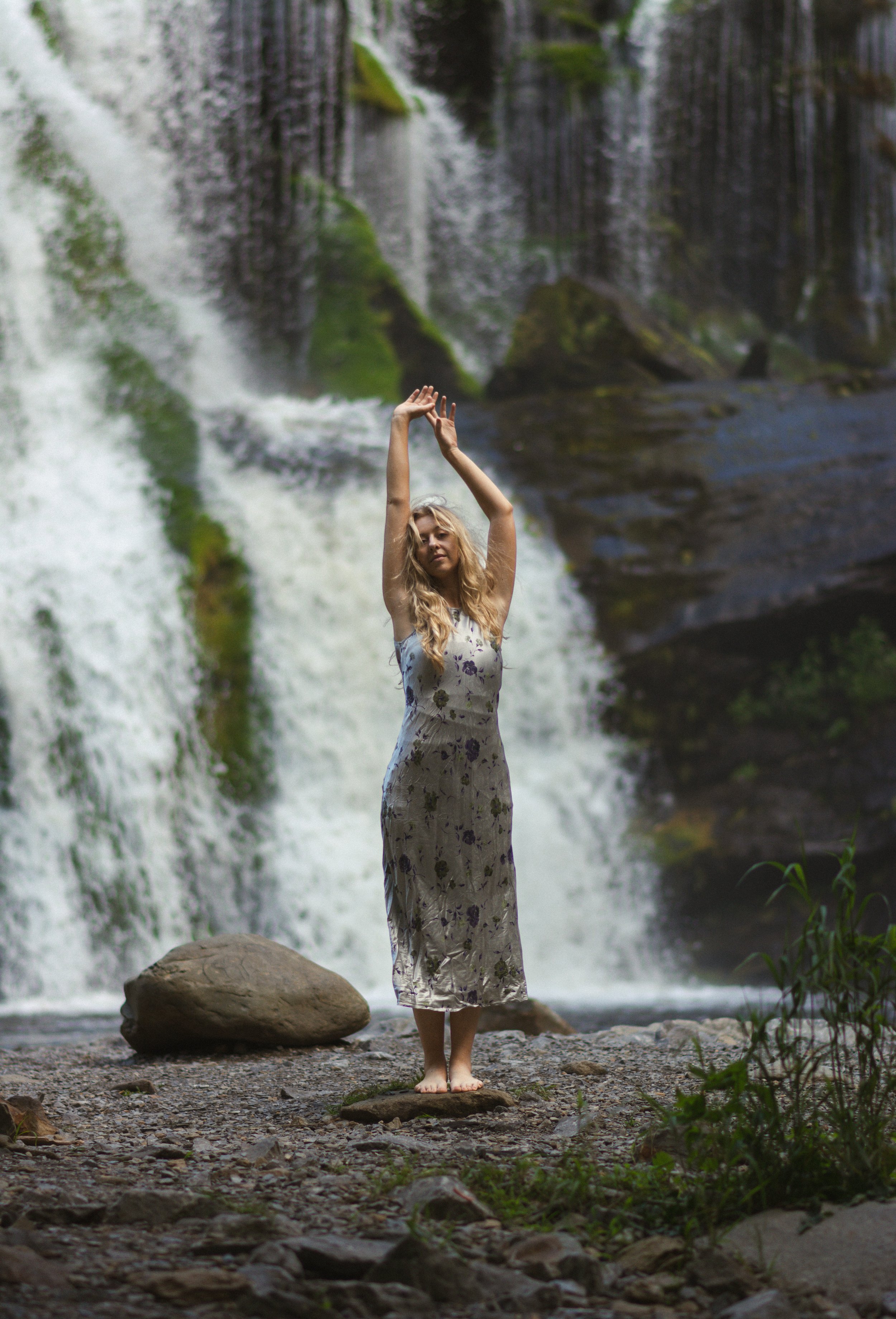 A woman with long blonde hair dancing with her arms raised in front of a waterfall in a natural outdoor setting.