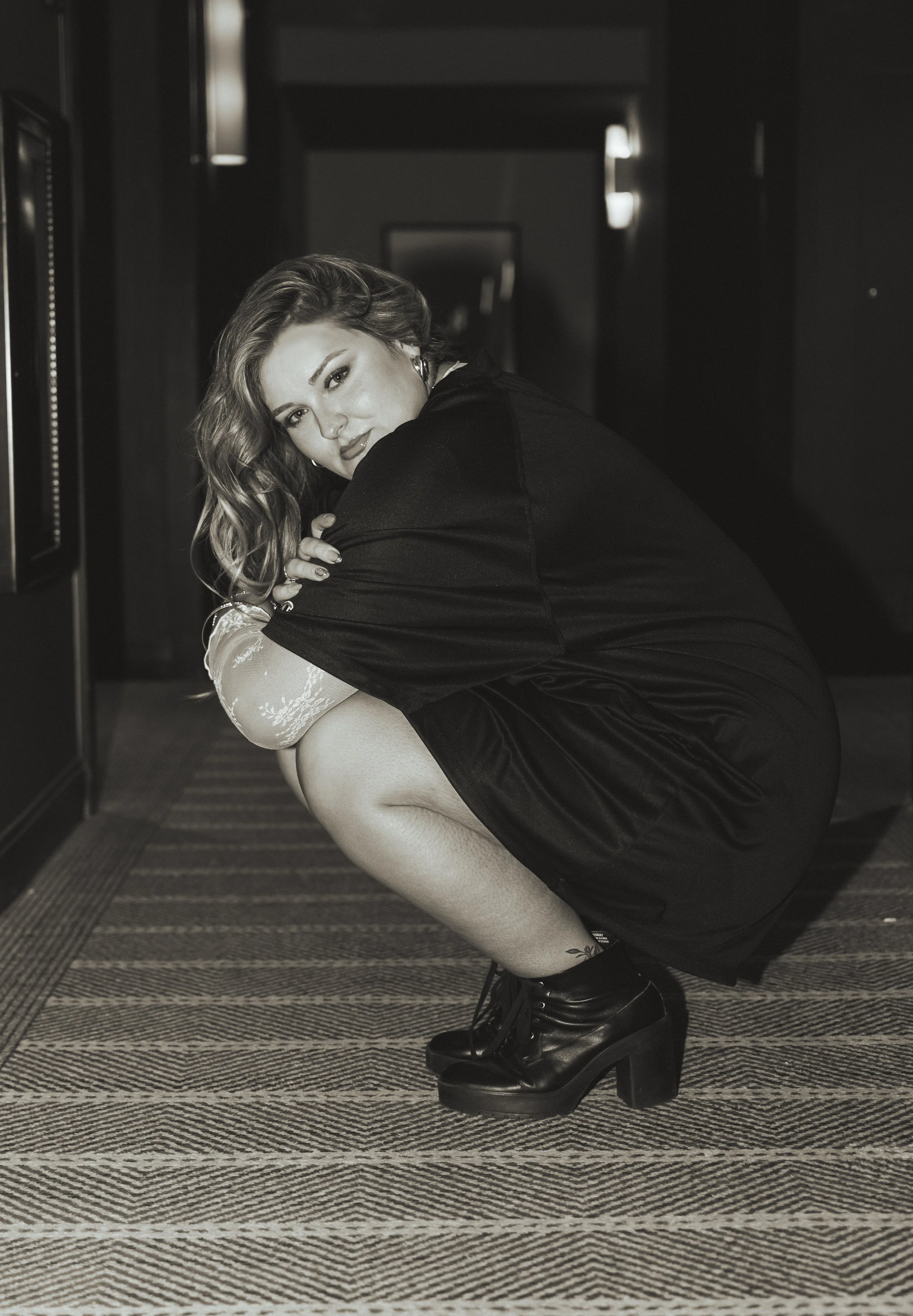 A woman with wavy hair and makeup leaning over, wearing a black top, fishnet stockings, and black platform boots, squatting on a patterned carpet in a dimly lit hallway with dark walls.