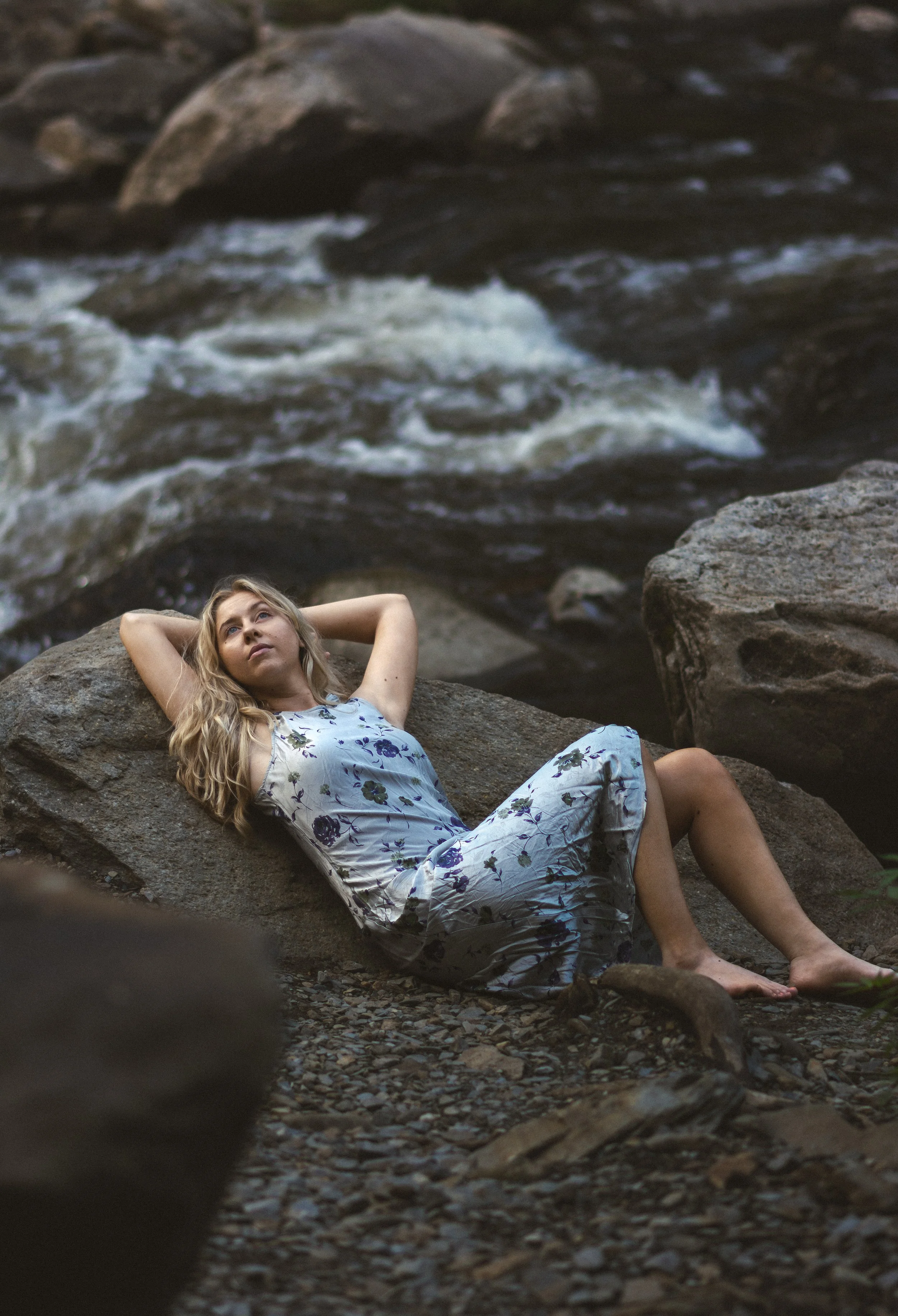 A young woman with long blonde hair relaxing on a large rock near a flowing river, wearing a floral dress.