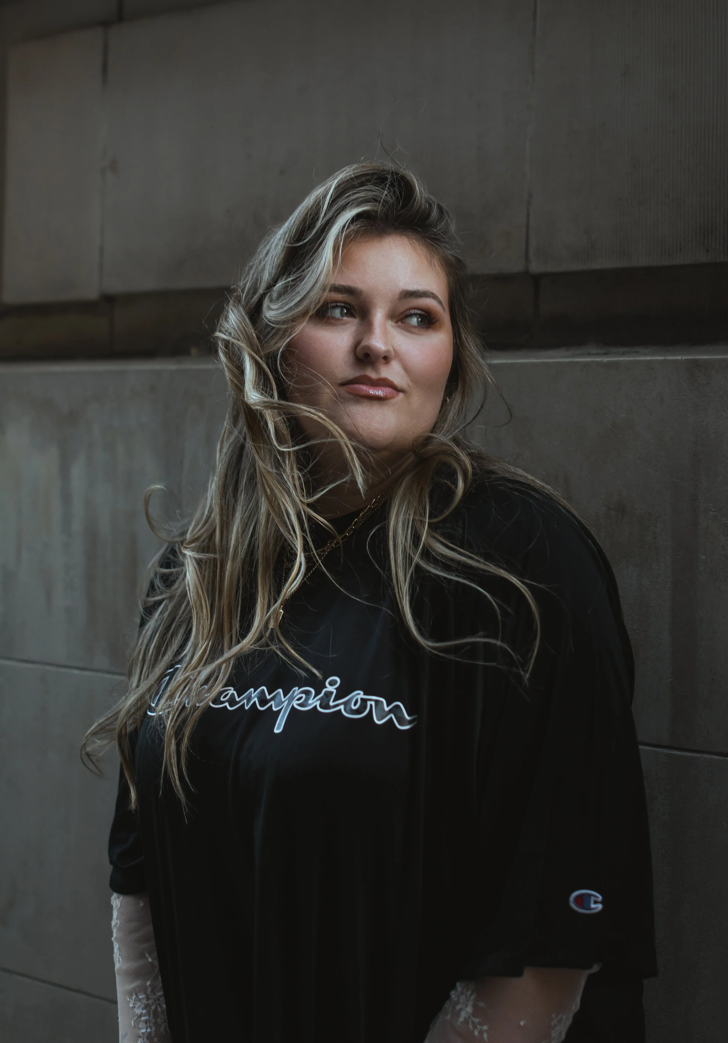 A young woman with long, curly blond hair and blue eyes, wearing a black sweatshirt with the word 'champion' on it, standing against a concrete wall.