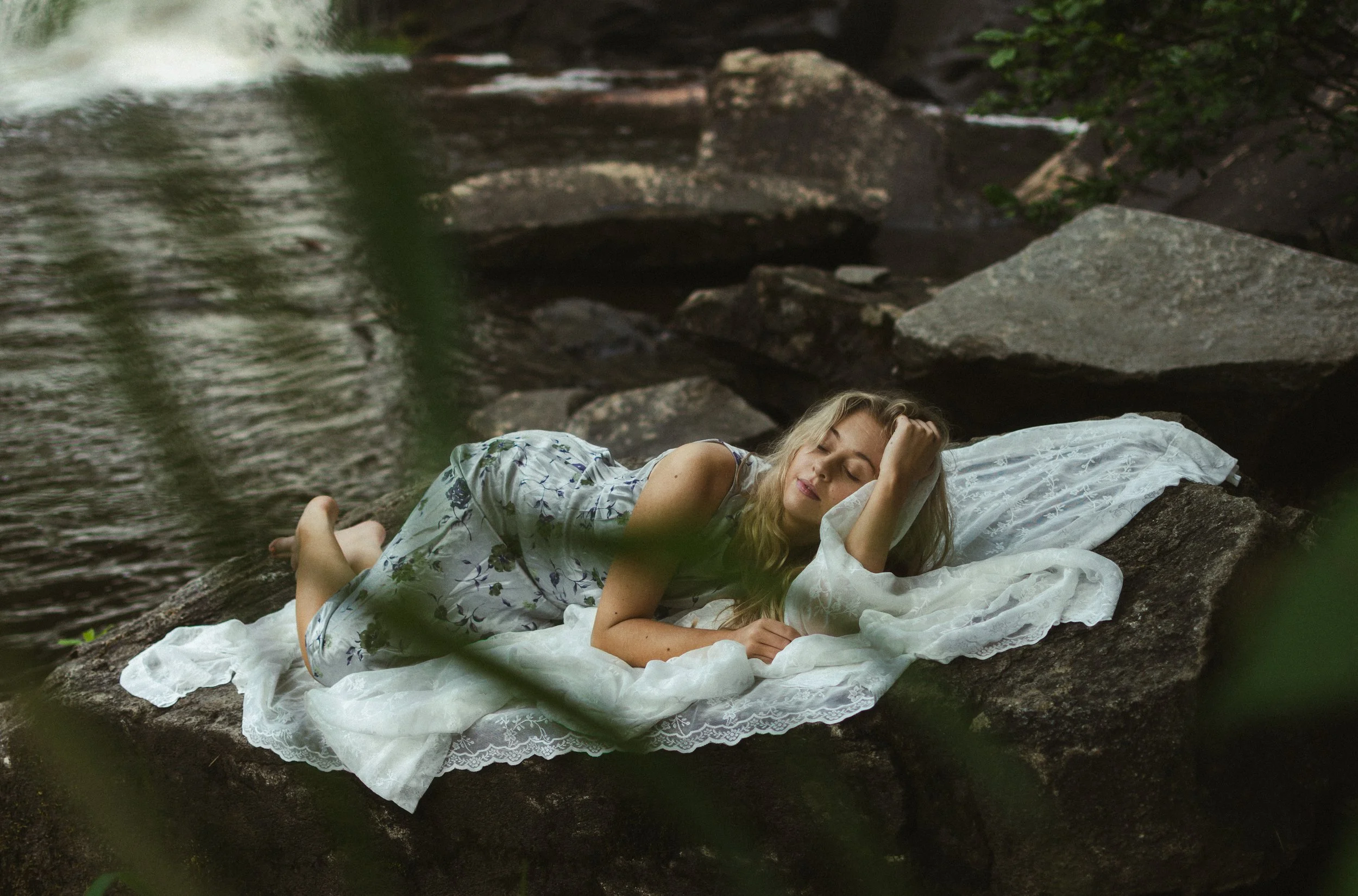 A woman with long hair lies peacefully on a white lace blanket on rocks near a stream in a forest. She has her eyes closed, resting her head on her hand.