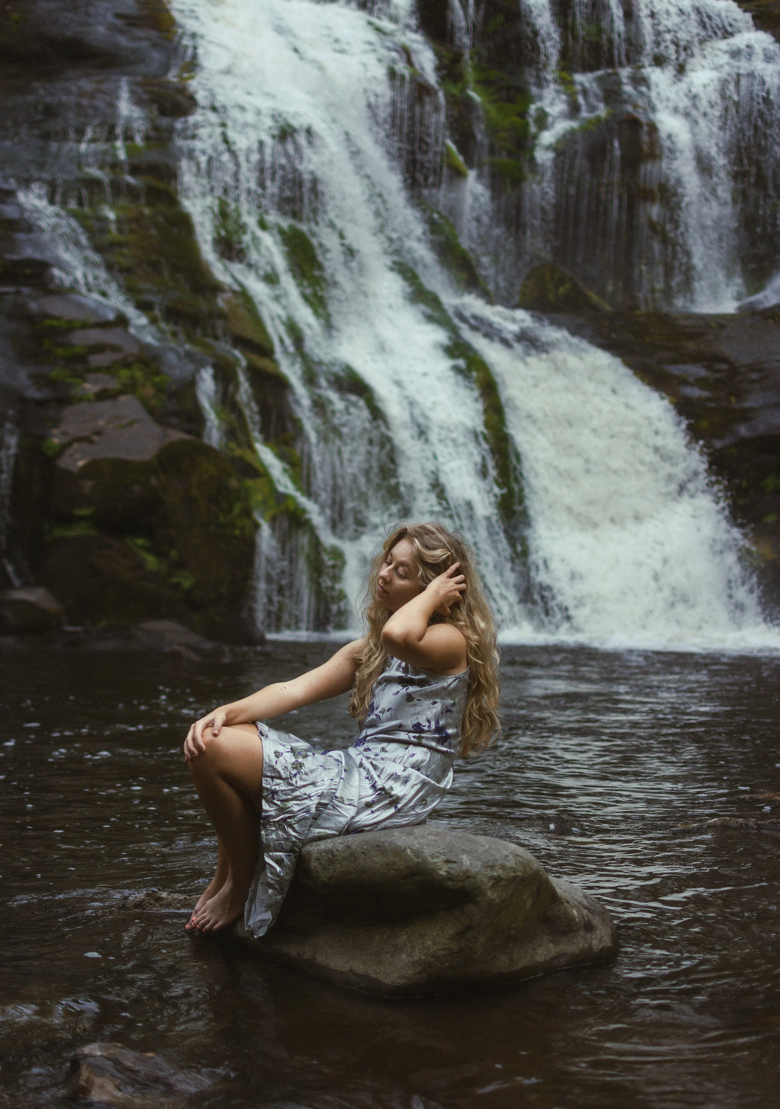 A woman with long blonde hair sitting on a large rock in a shallow river, with a waterfall in the background.