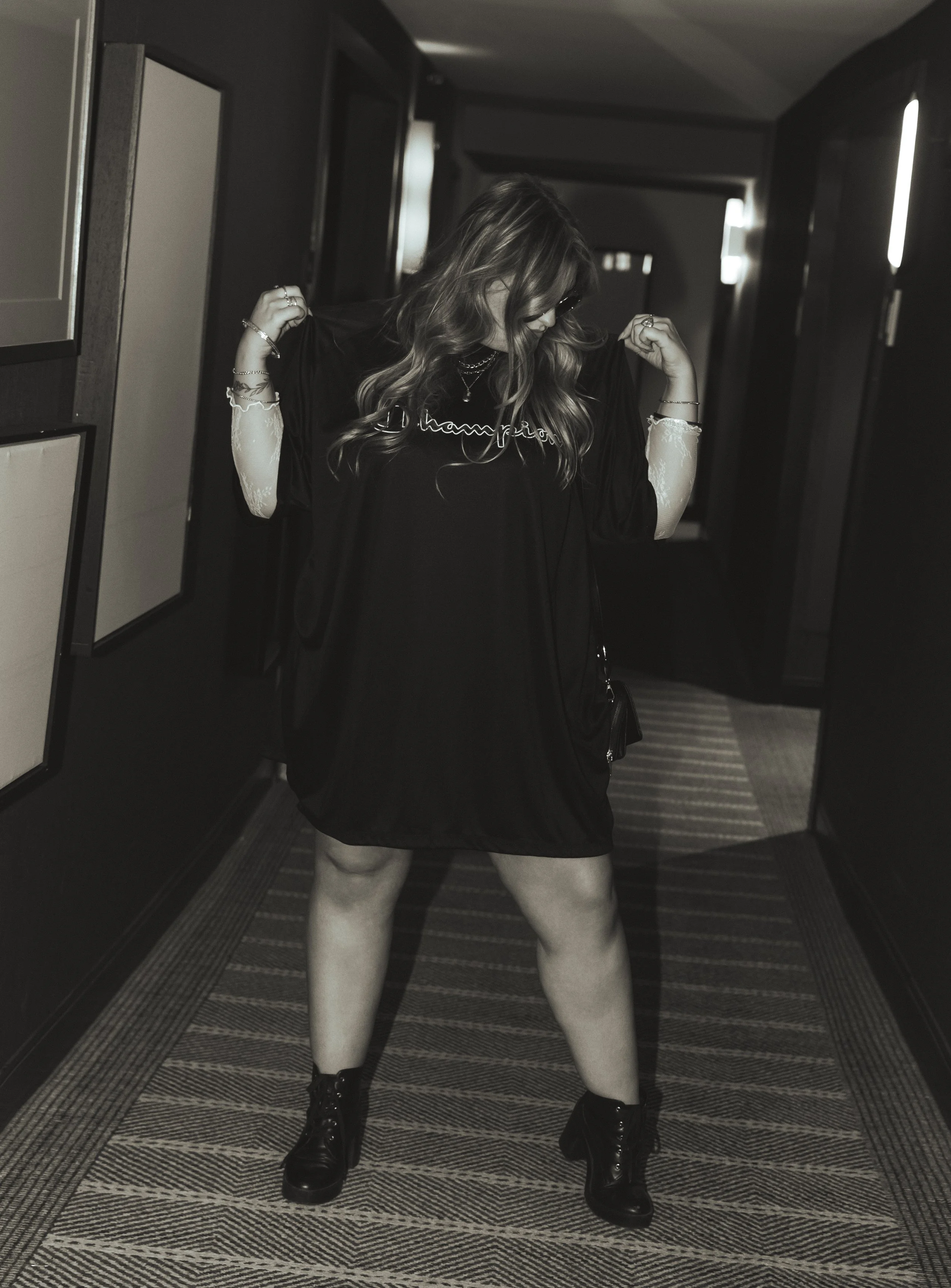 A woman with wavy hair, wearing dark sunglasses, a black and white long-sleeve shirt under a black graphic t-shirt, a necklace, bracelets, and black boots, standing in an indoor corridor with dark wooden walls and striped carpet, flexing her arms.