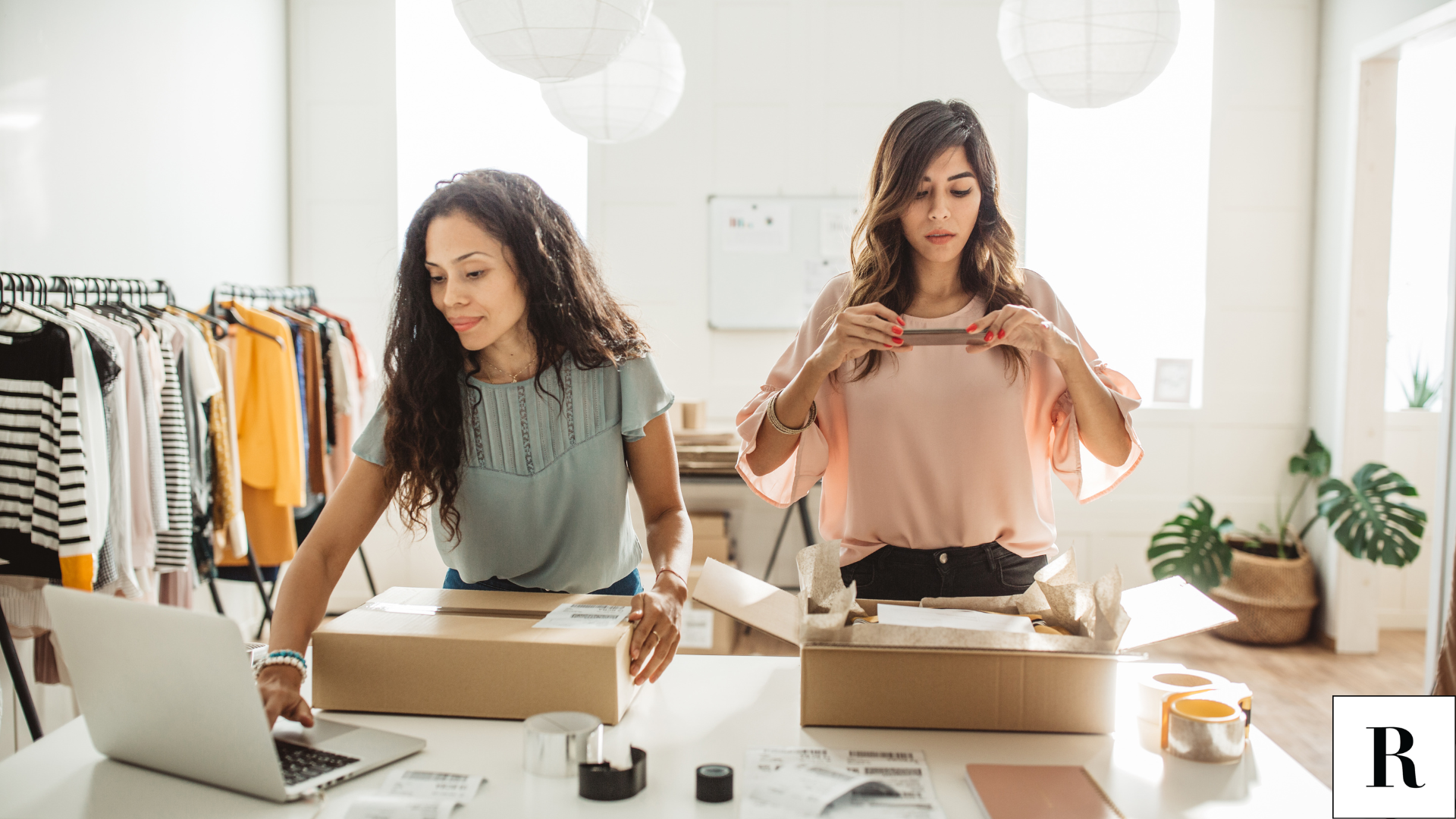 two female business owners packing up shipments for their customers. Racks of clothing in the background