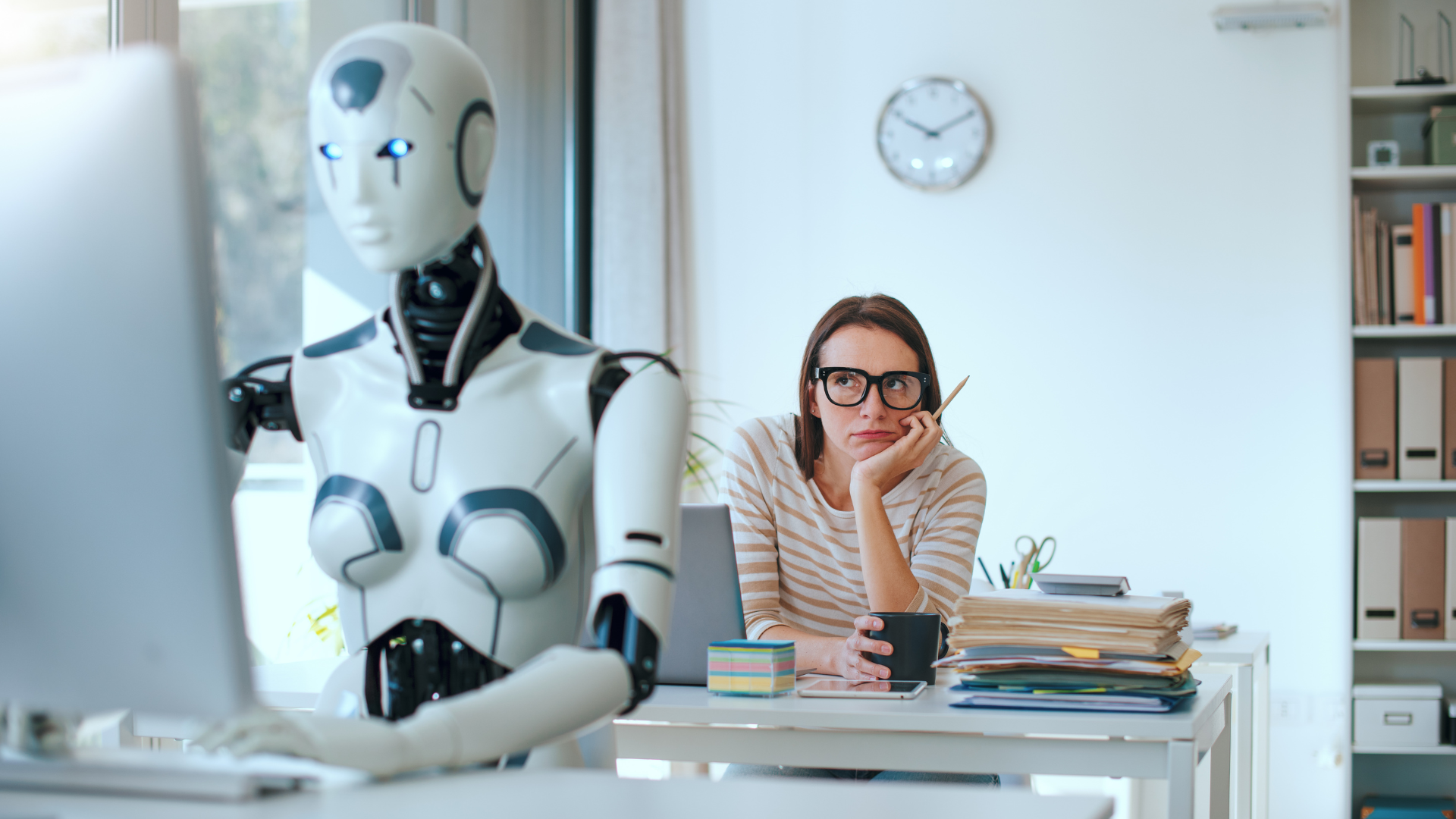 a woman sitting at her desk in an office. She is staring at the desk in front of her with frustration. The desk in front of her is occupied by a robot working at their computer.