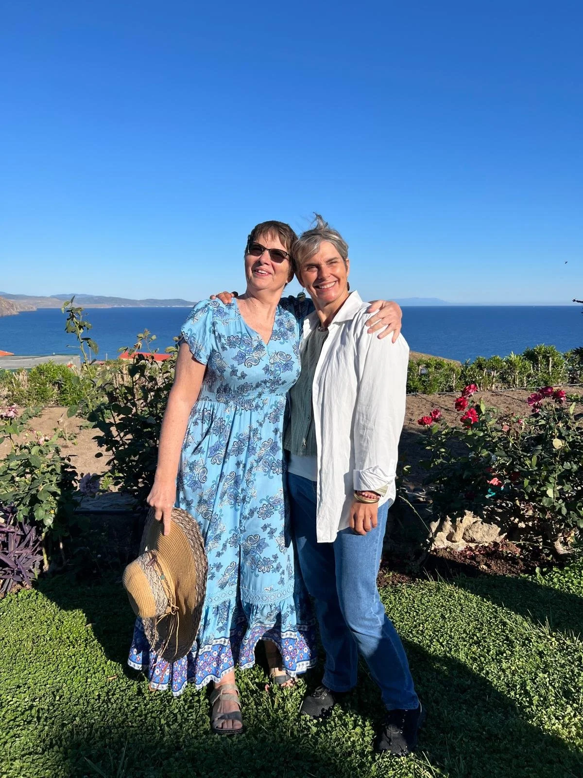 two women smiling at the edge of a garden, with the sea and blue sky behind them