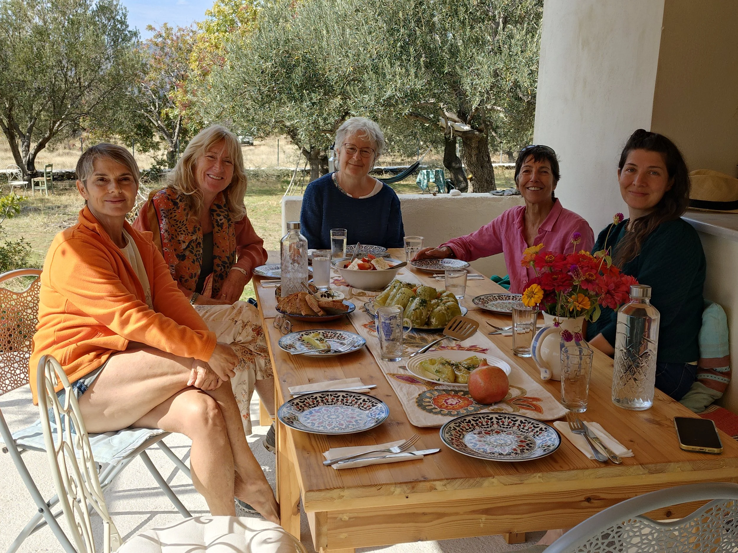 five middle aged women sitting around a table in an olive grove, smiling into the camera just as they are about to have lunch.